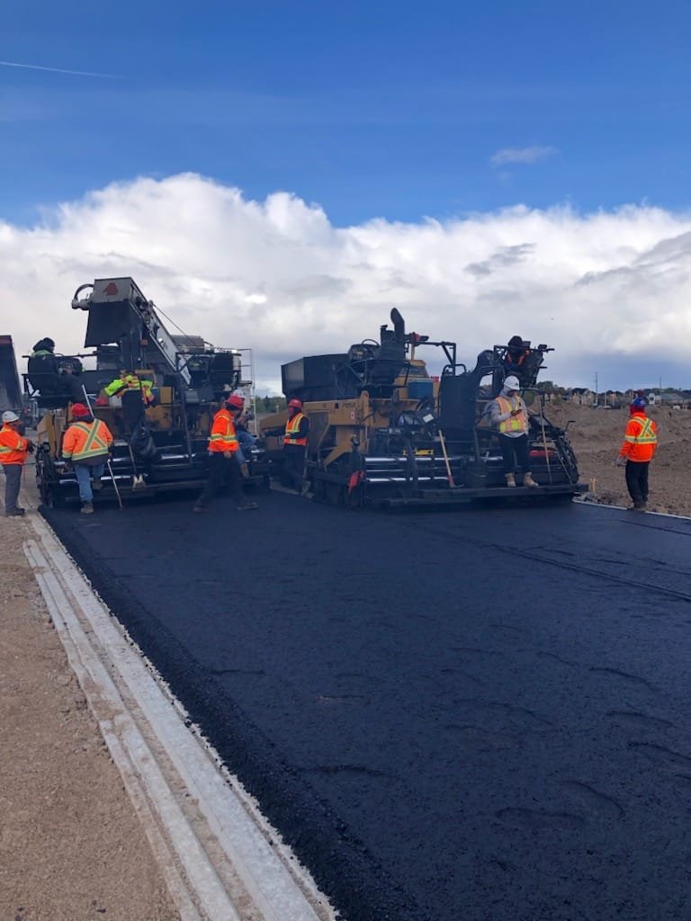 Workers in orange safety vests and helmets paving a road with asphalt using industrial paving machines, under a partly cloudy sky.