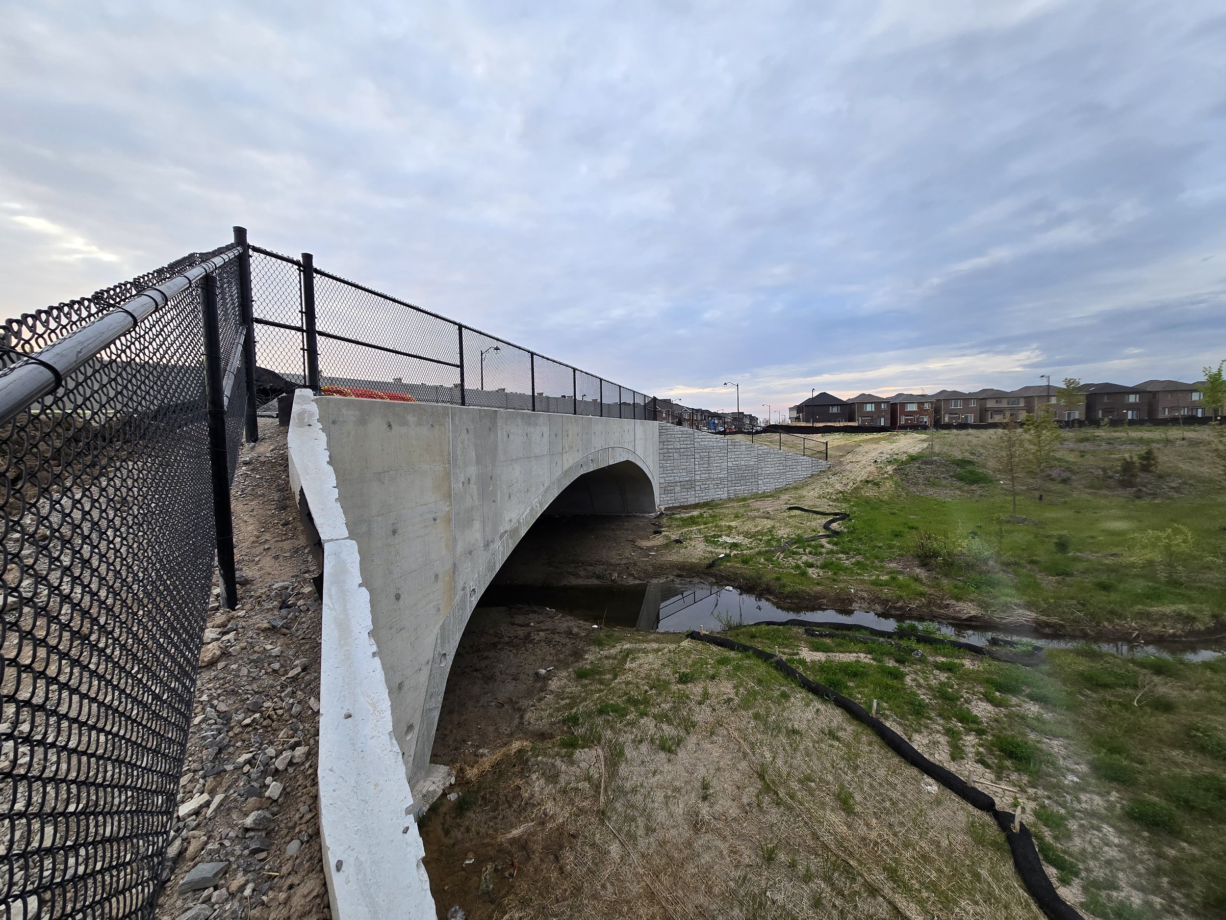View of a small bridge over a creek in a residential area, with houses visible in the background and a cloudy sky overhead.