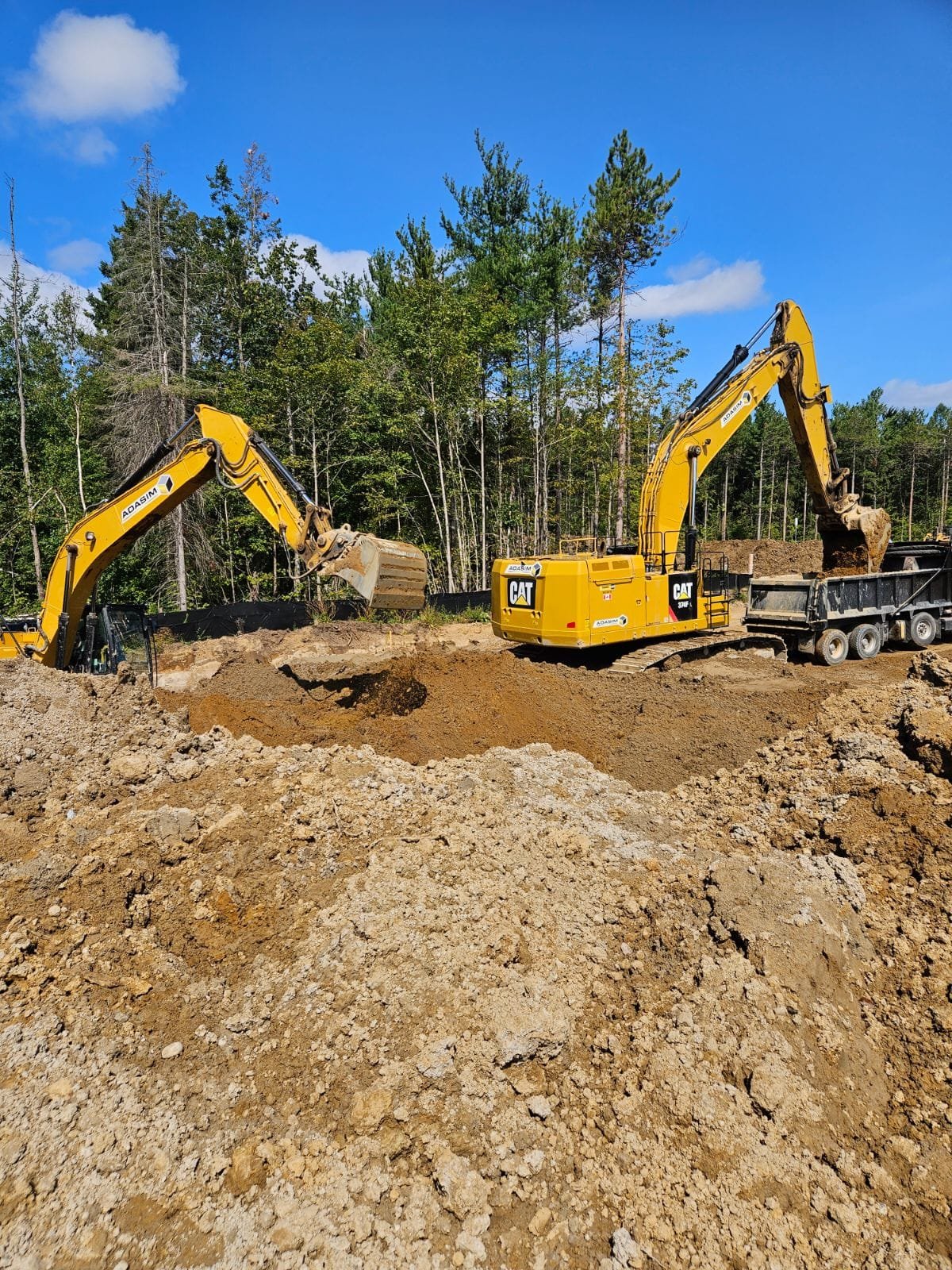 Two yellow excavators are moving dirt at a construction site with a background of trees and a blue sky with clouds.