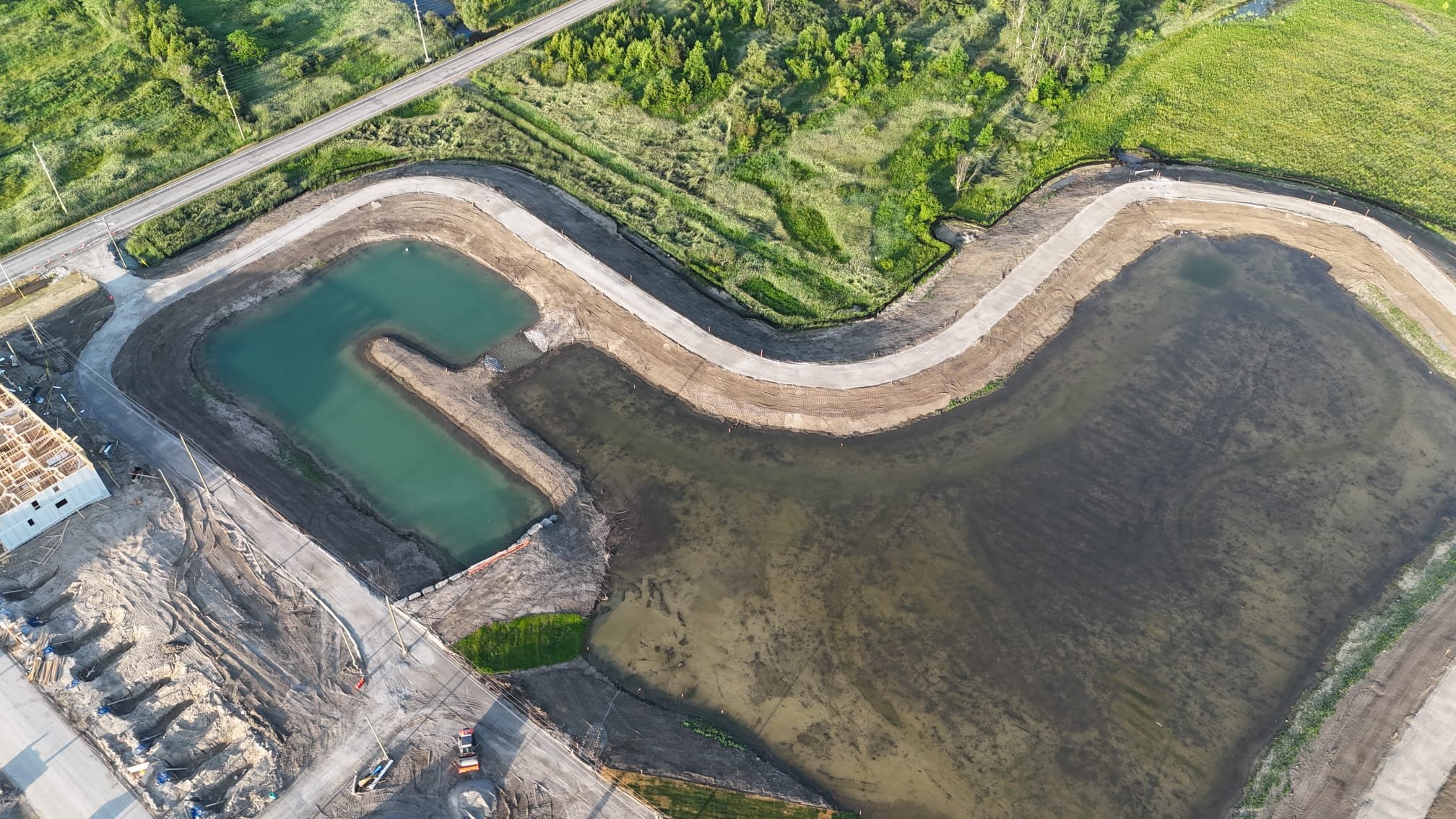 Aerial view of a construction site for a new water body, with the partially built pond or lake, surrounded by dirt and construction materials, adjacent to a green grassy area and a road.