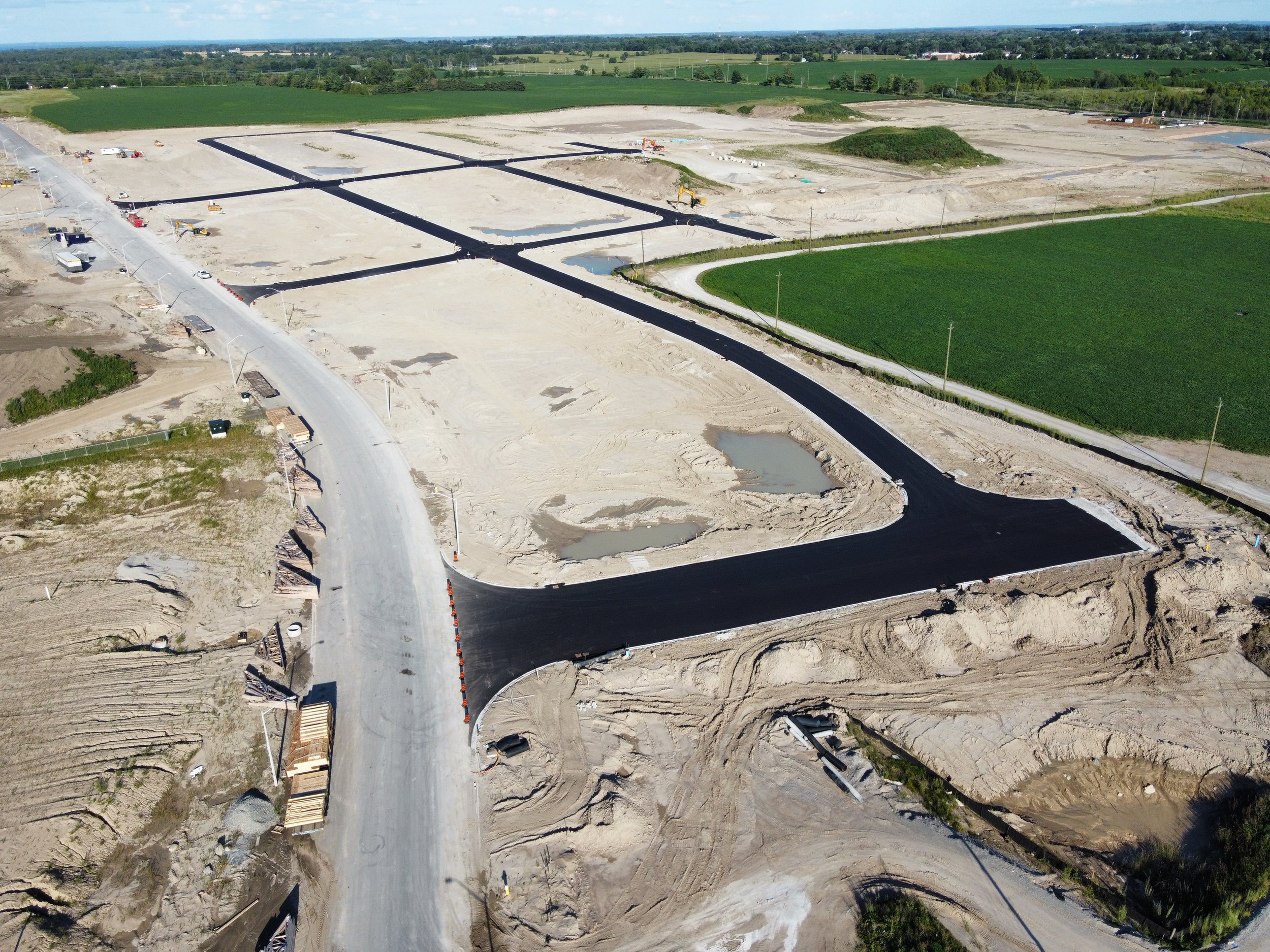 Aerial view of a construction site with asphalt paving, unfinished roads, and surrounding farmland in the distance.
