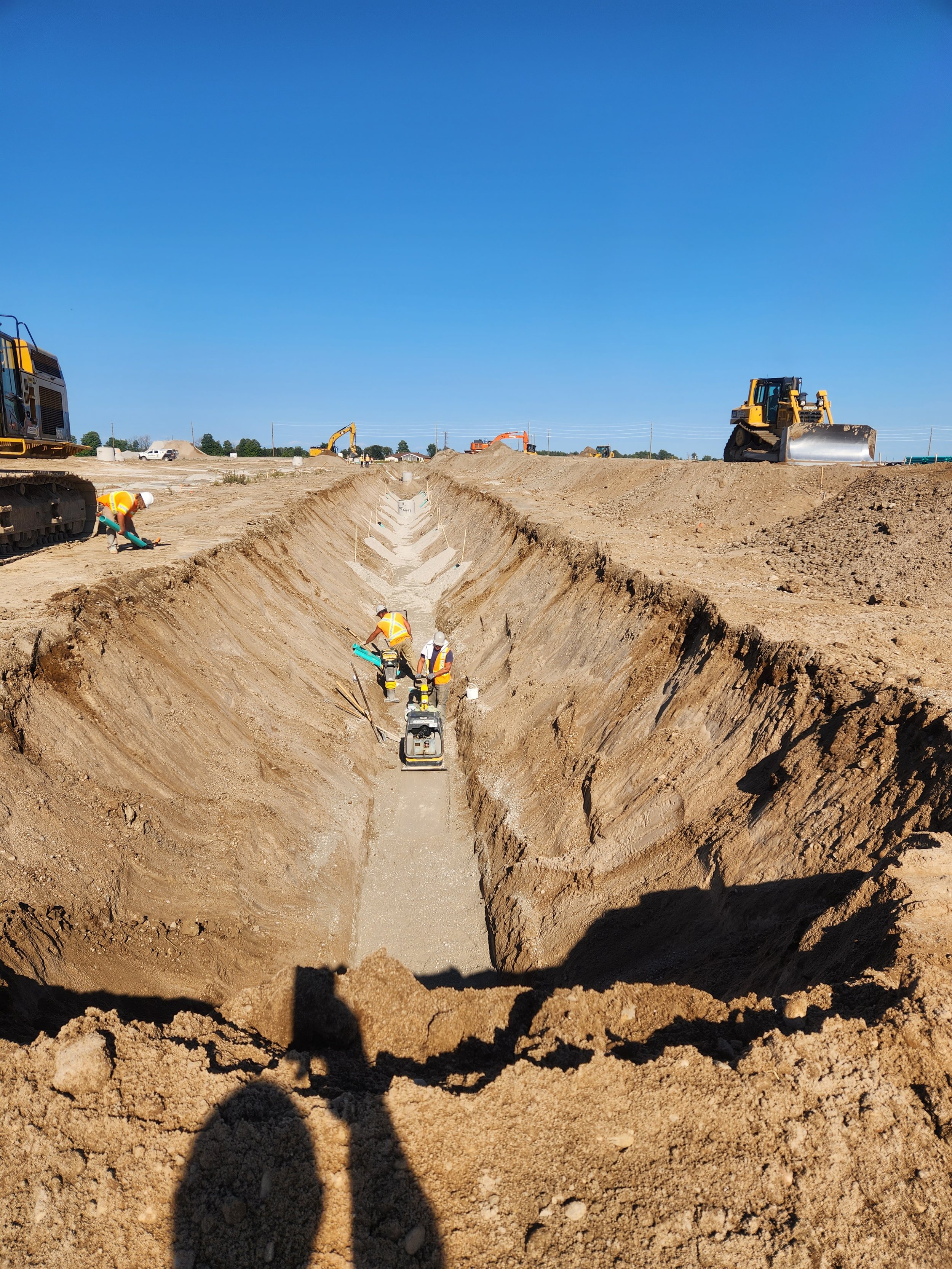 Construction workers and heavy machinery at a construction site with a deep trench and clear blue sky.