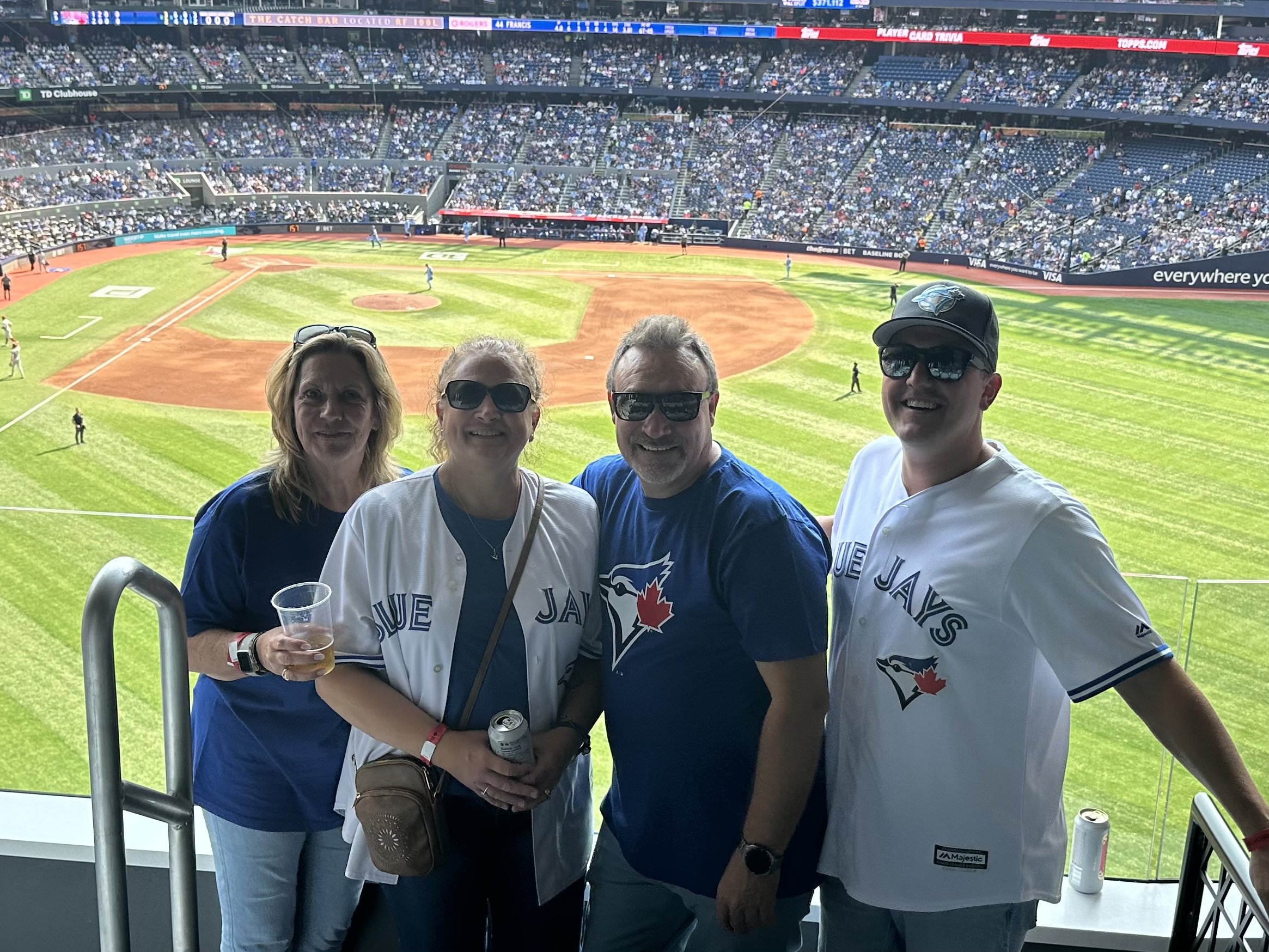 Four people standing in the stadium seats overlooking the baseball field during a game. They are wearing Toronto Blue Jays jerseys and sunglasses, with a mostly empty stadium background.