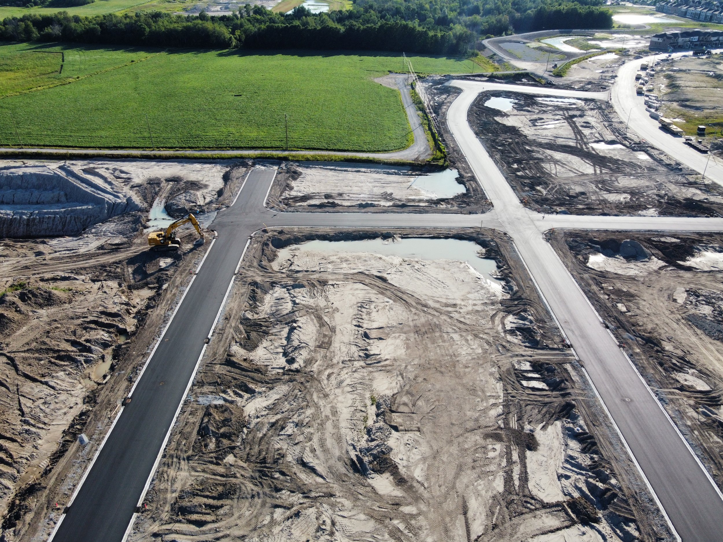 An aerial view of a construction site with roads being built, with some sections of roads paved and others still under construction, and a yellow excavator working on the ground.