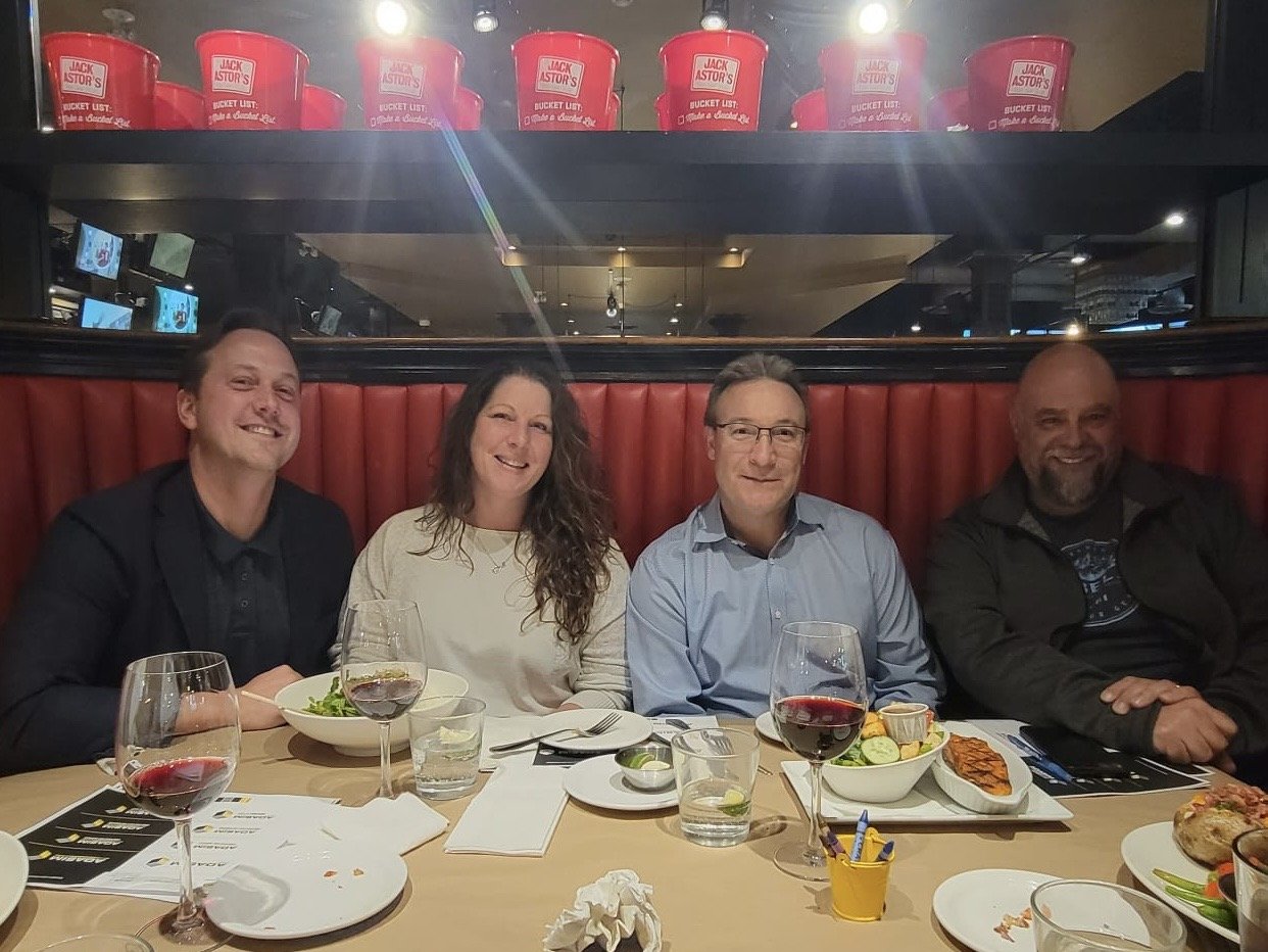 Four people sitting at a restaurant table with food and drinks, smiling. Behind them, a red wall with a mirror showing a reflection of the restaurant interior. On the table, there are glasses, plates, and a small yellow cup with pens.