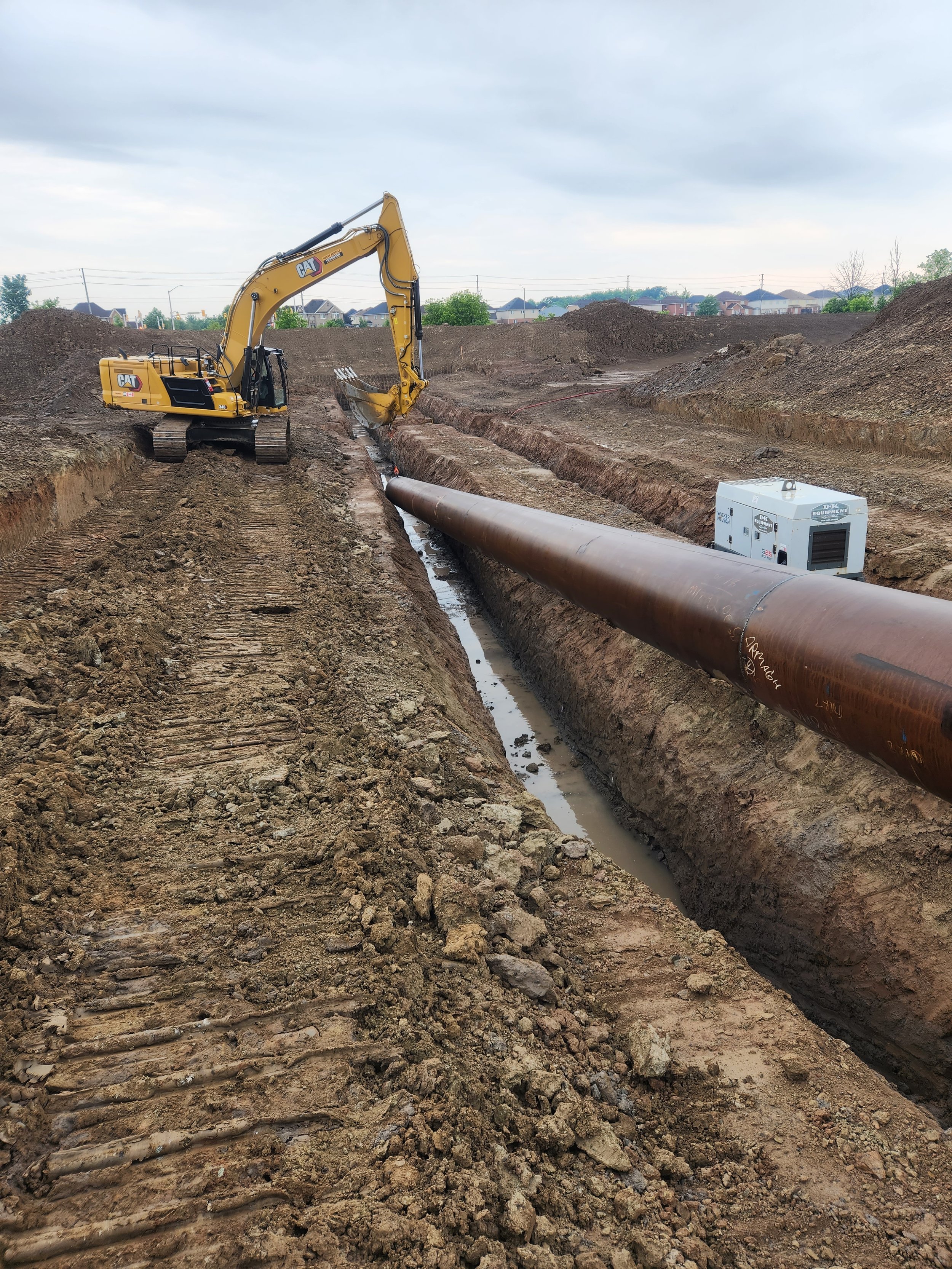 Construction site with a large excavator working on underground pipe installation, with dirt and soil excavated into trenches and a generator nearby.
