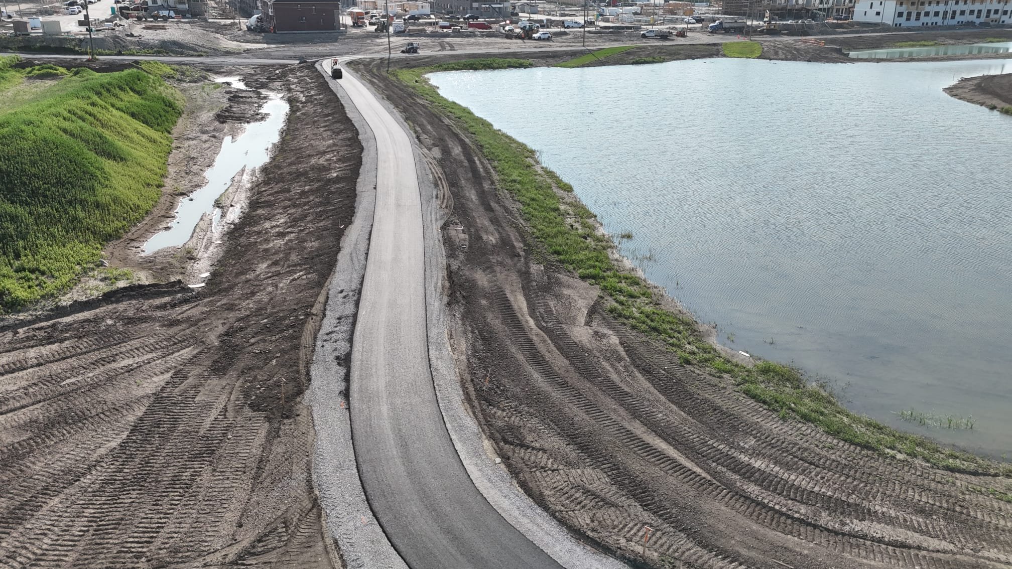 A construction site with a newly paved road bordering a river, with earthwork and machinery tracks visible on the sides of the road, and a parking lot with cars in the background.
