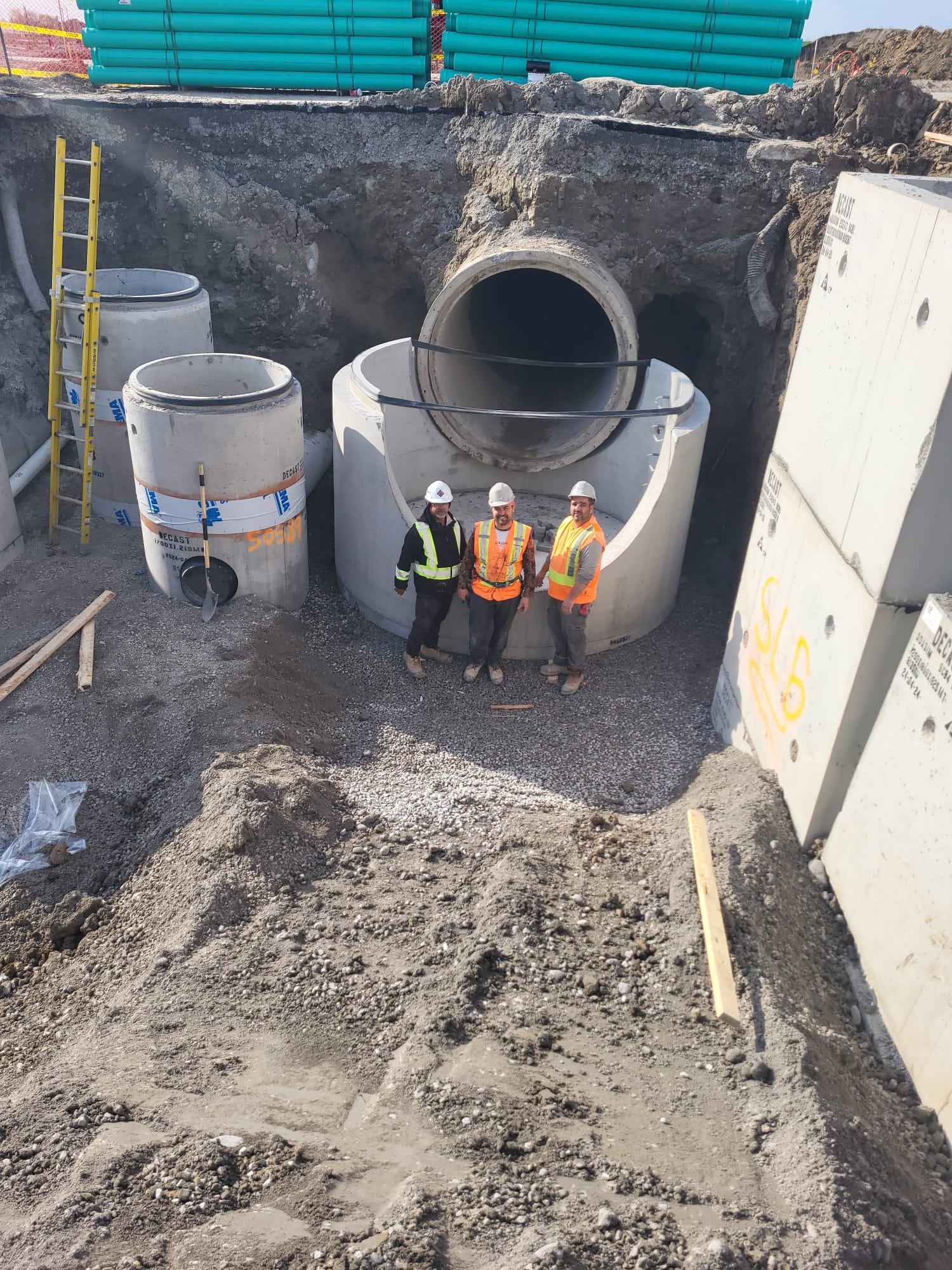 Three construction workers wearing safety vests and helmets standing in front of large concrete pipe segments at a construction site.