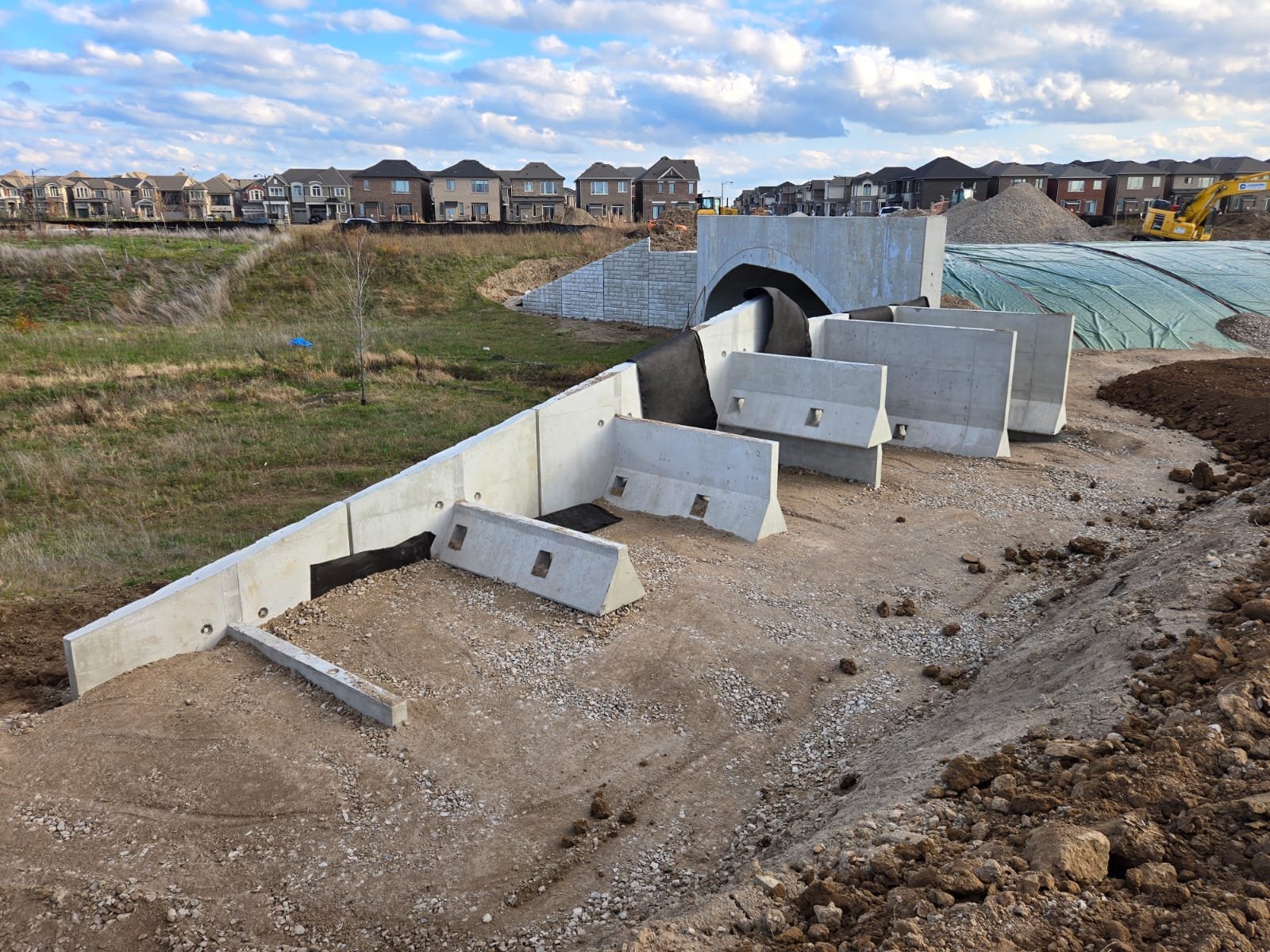 Construction site with concrete barriers and a tunnel under construction, residential houses in the background, blue sky with clouds.