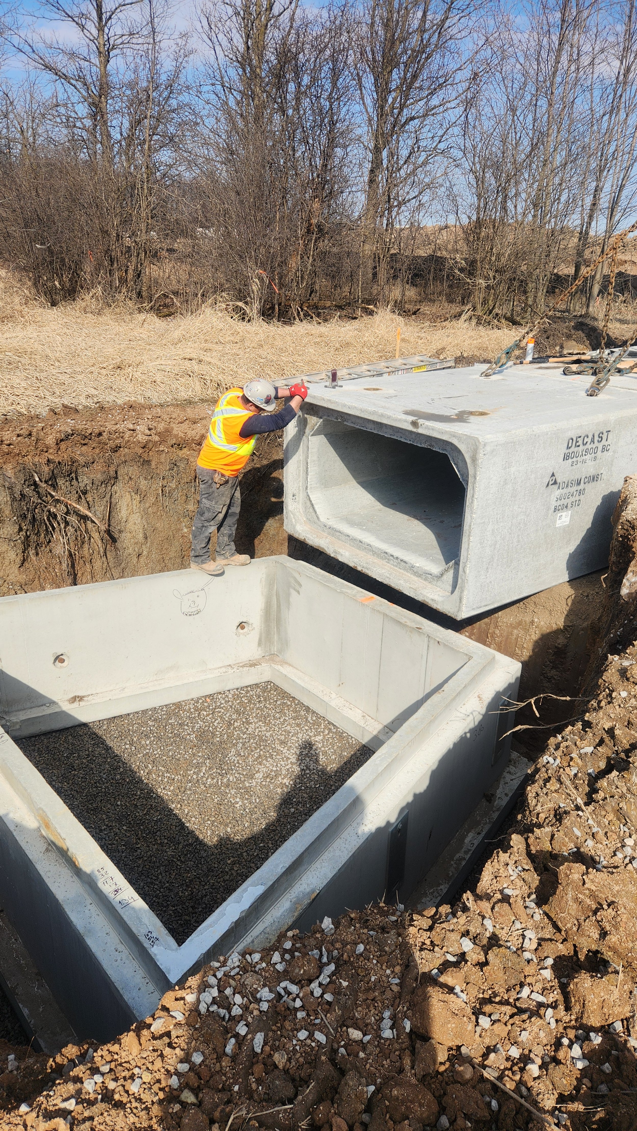Construction worker installing concrete culvert underground at construction site with trees and dirt in background.