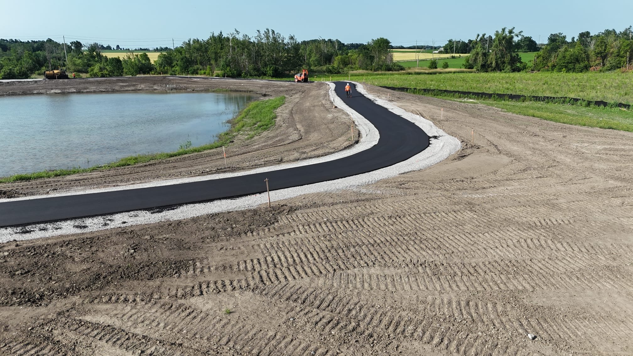 Construction workers paving a winding asphalt road by a pond with lush greenery and fields in the background.