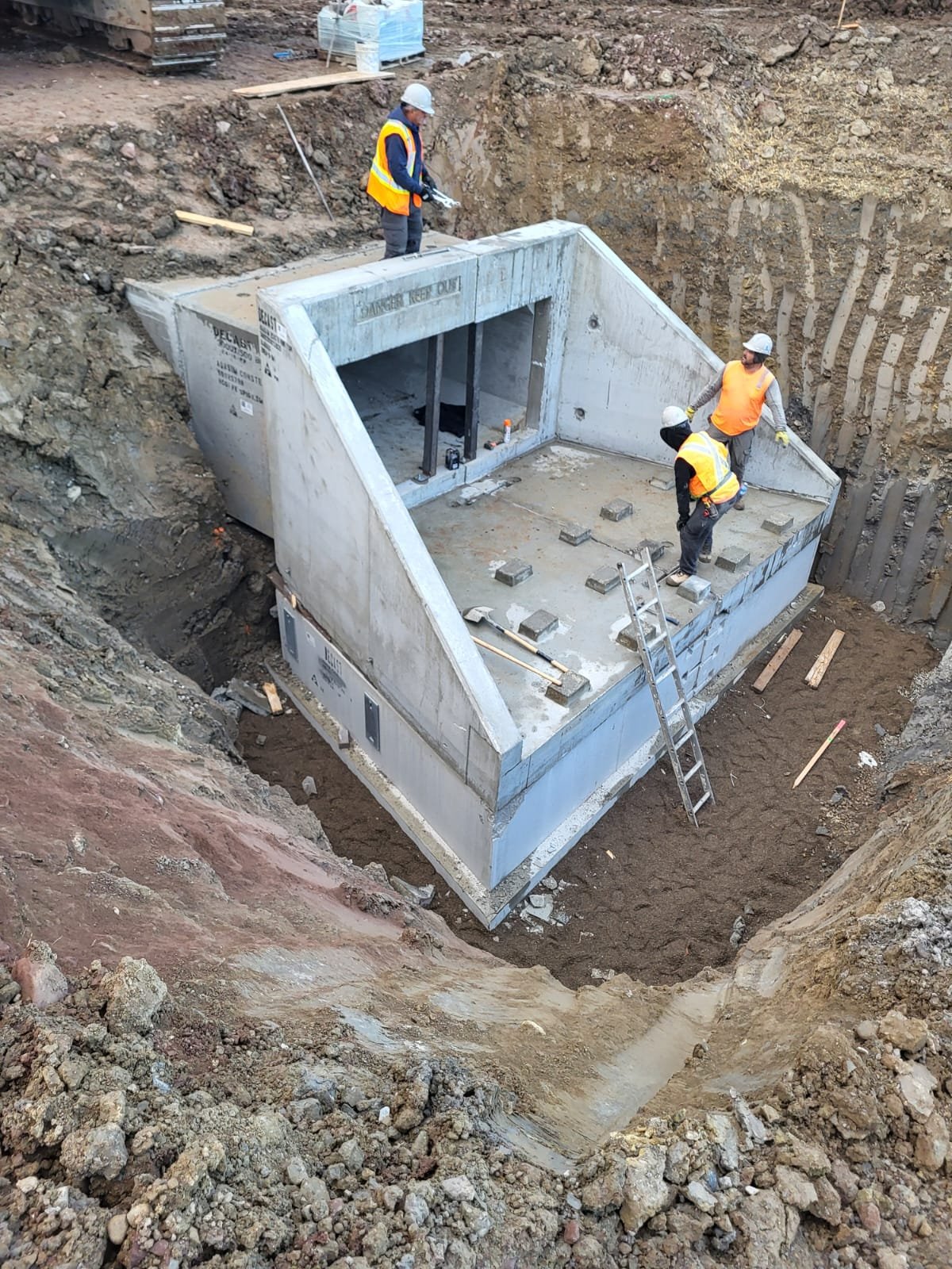 Construction workers install a concrete structure in a deep excavation site with steel reinforcements visible inside.
