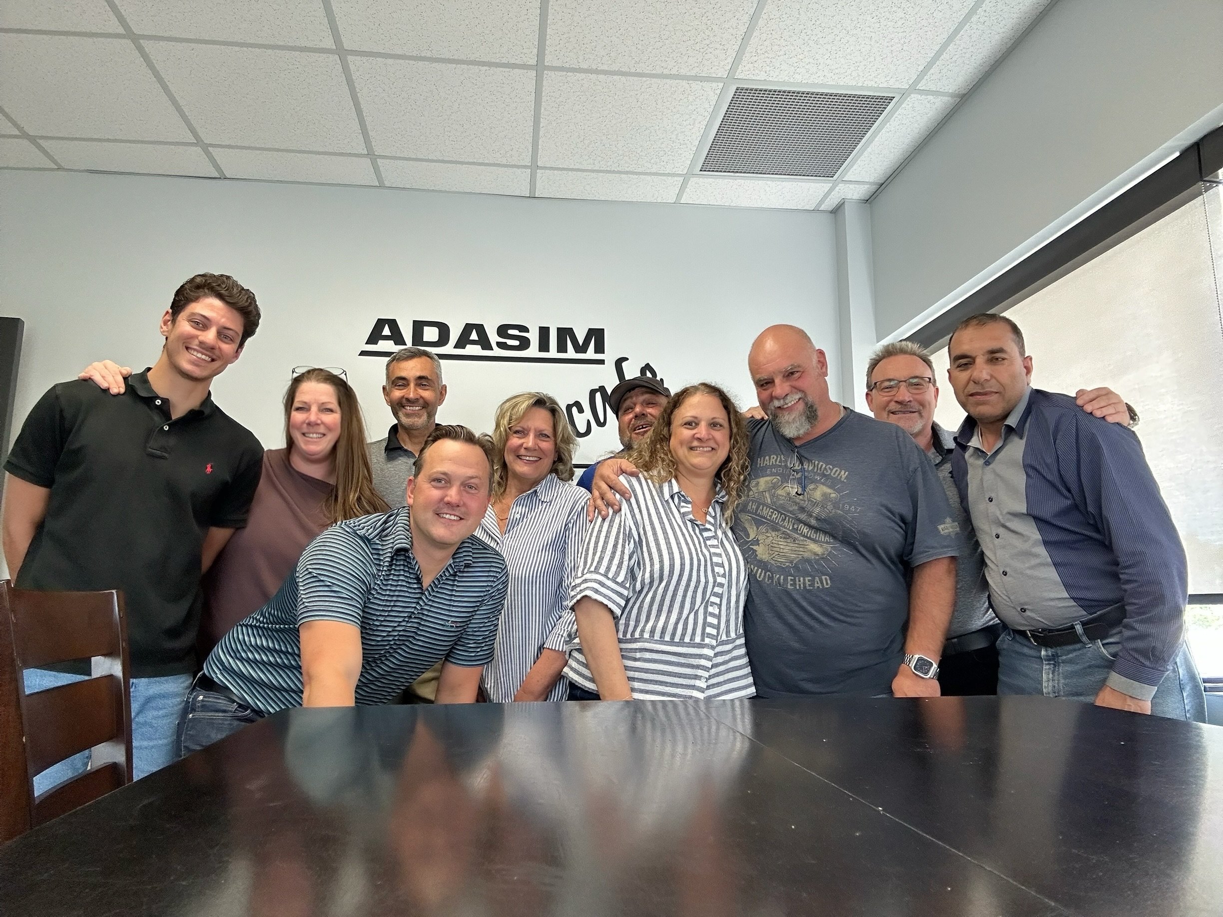 Group of ten people standing together indoors, smiling, in front of a wall with the words 'ADASIM' and some other text.