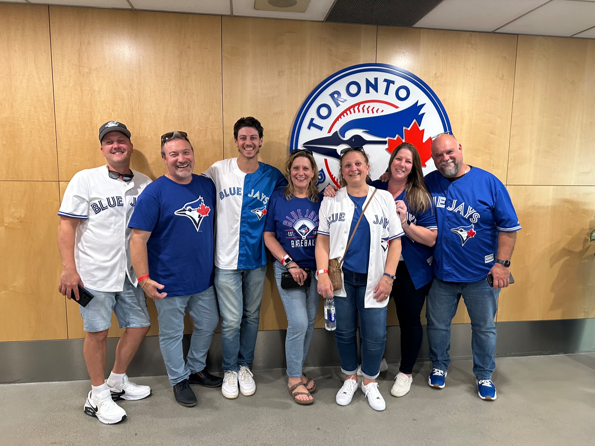 Group of seven people wearing Toronto Blue Jays baseball jerseys and shirts, standing in front of a large Toronto Blue Jays logo on a wall.