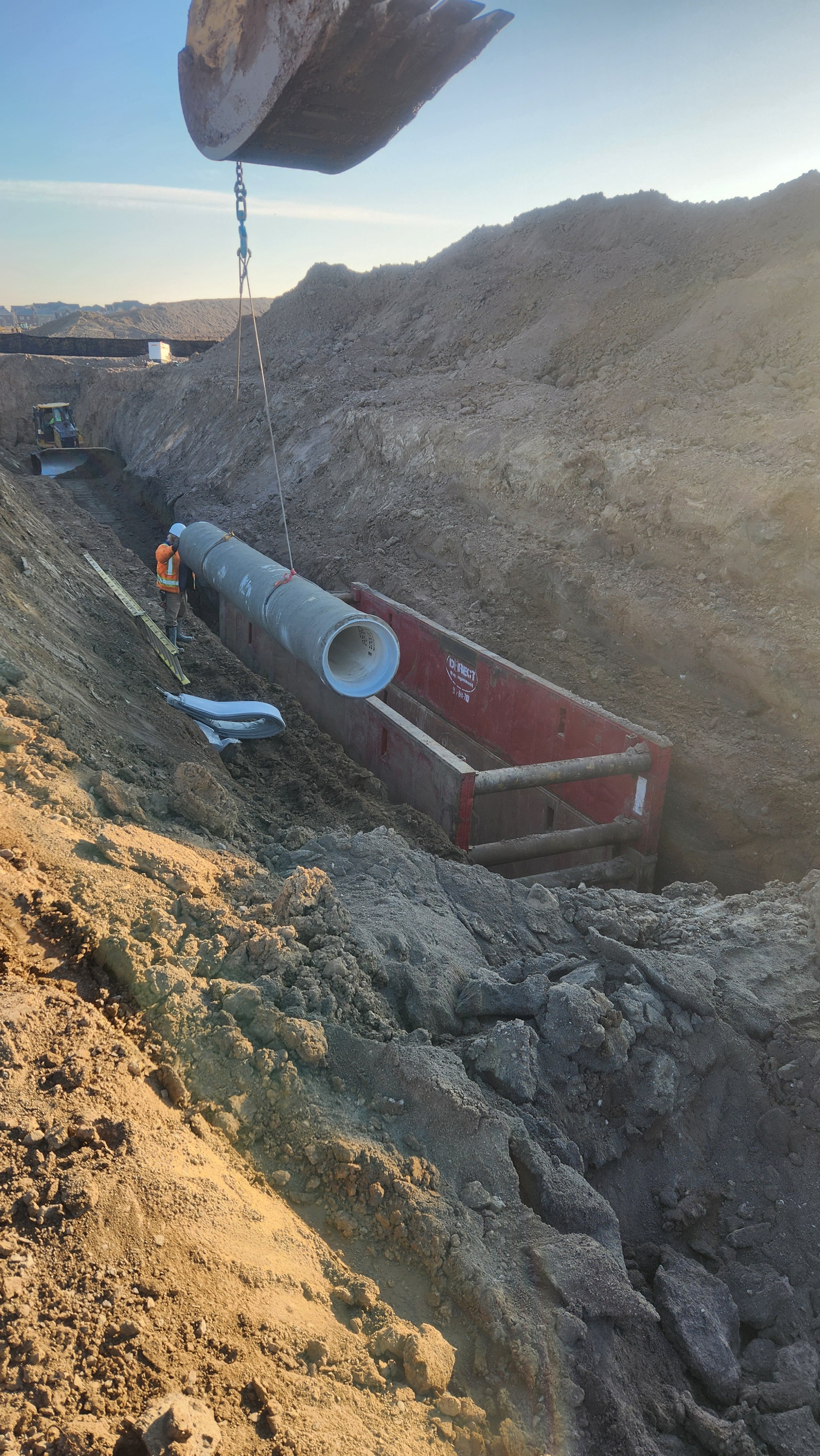 Construction workers installing a large pipe in a trench with machinery and dirt piles around.