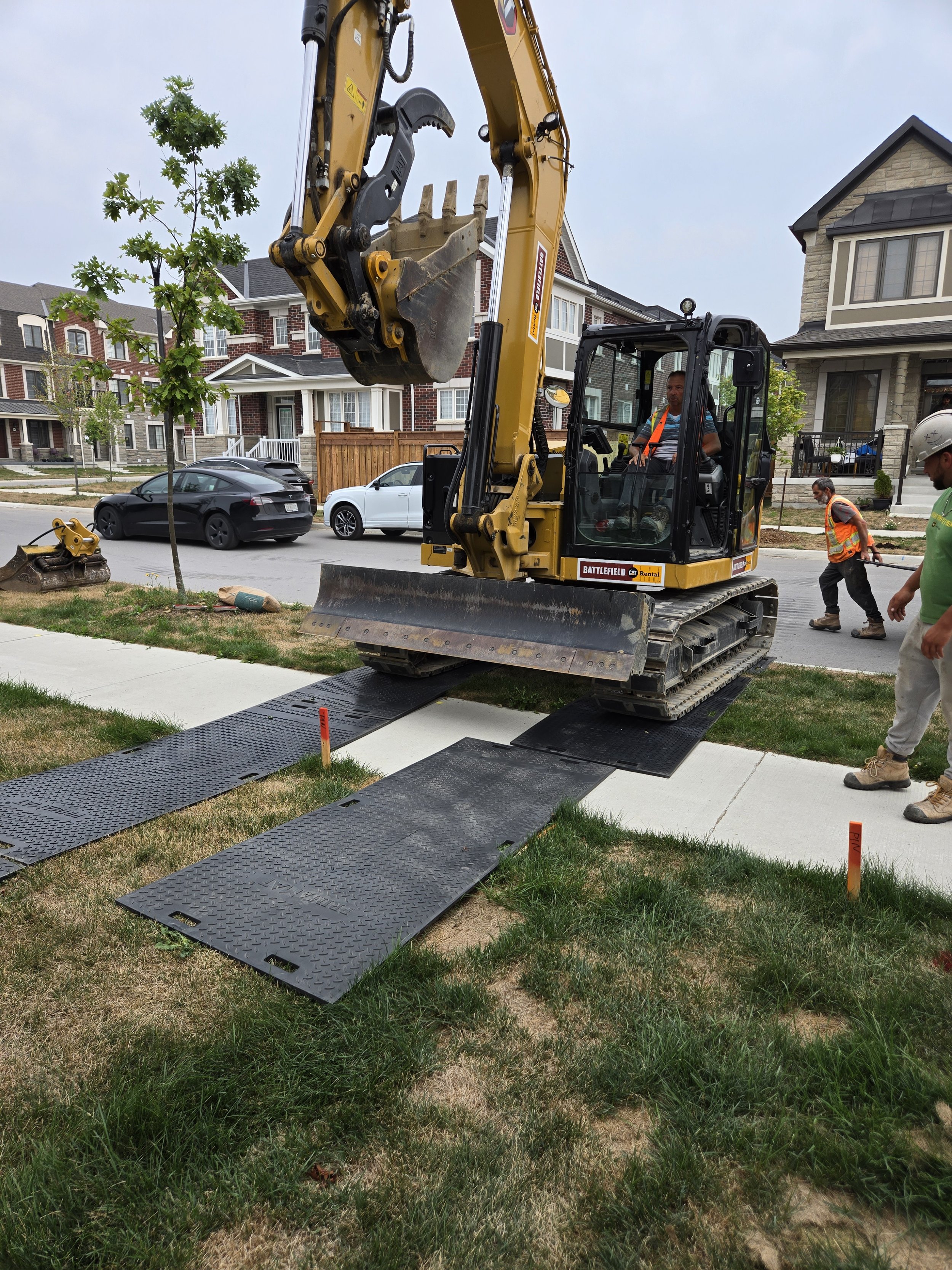 Construction workers operating a small excavator on a sidewalk, lifting a black ramp to assist with accessibility in a residential neighborhood.