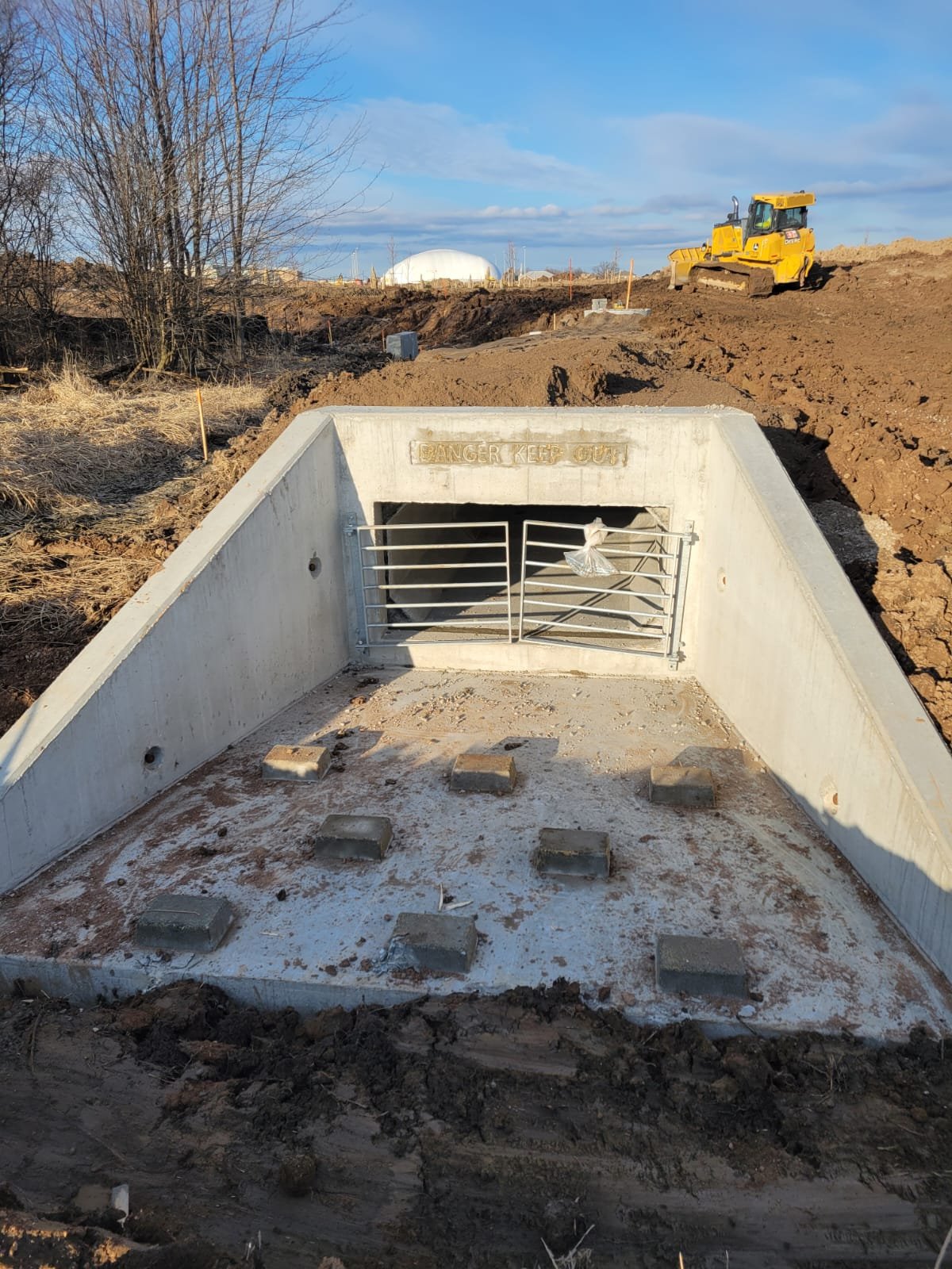 A concrete storm drain with a metal gate and a warning sign 'DANGER KEEP OUT.' Construction site with dirt and a yellow bulldozer in the background, under a blue sky with clouds.