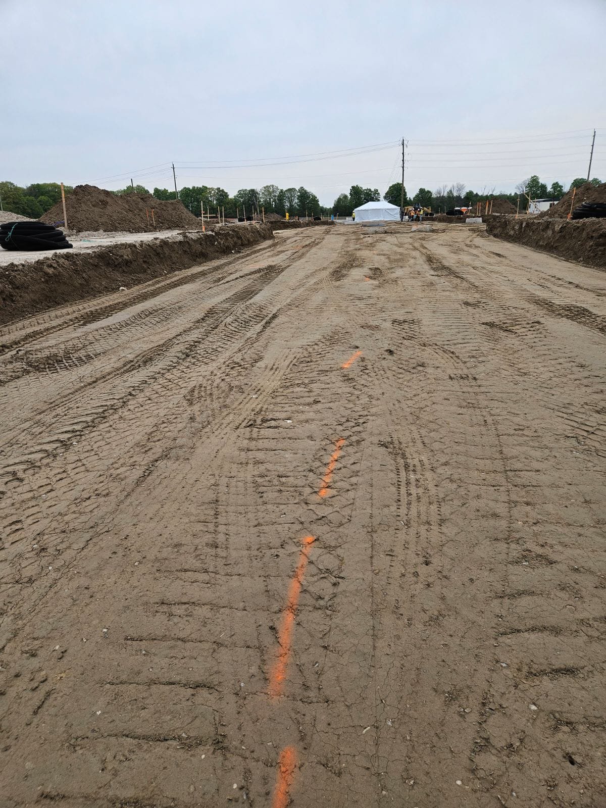 Construction site showing a dirt road with orange markings, piles of soil on the sides, and construction equipment in the background under a cloudy sky.