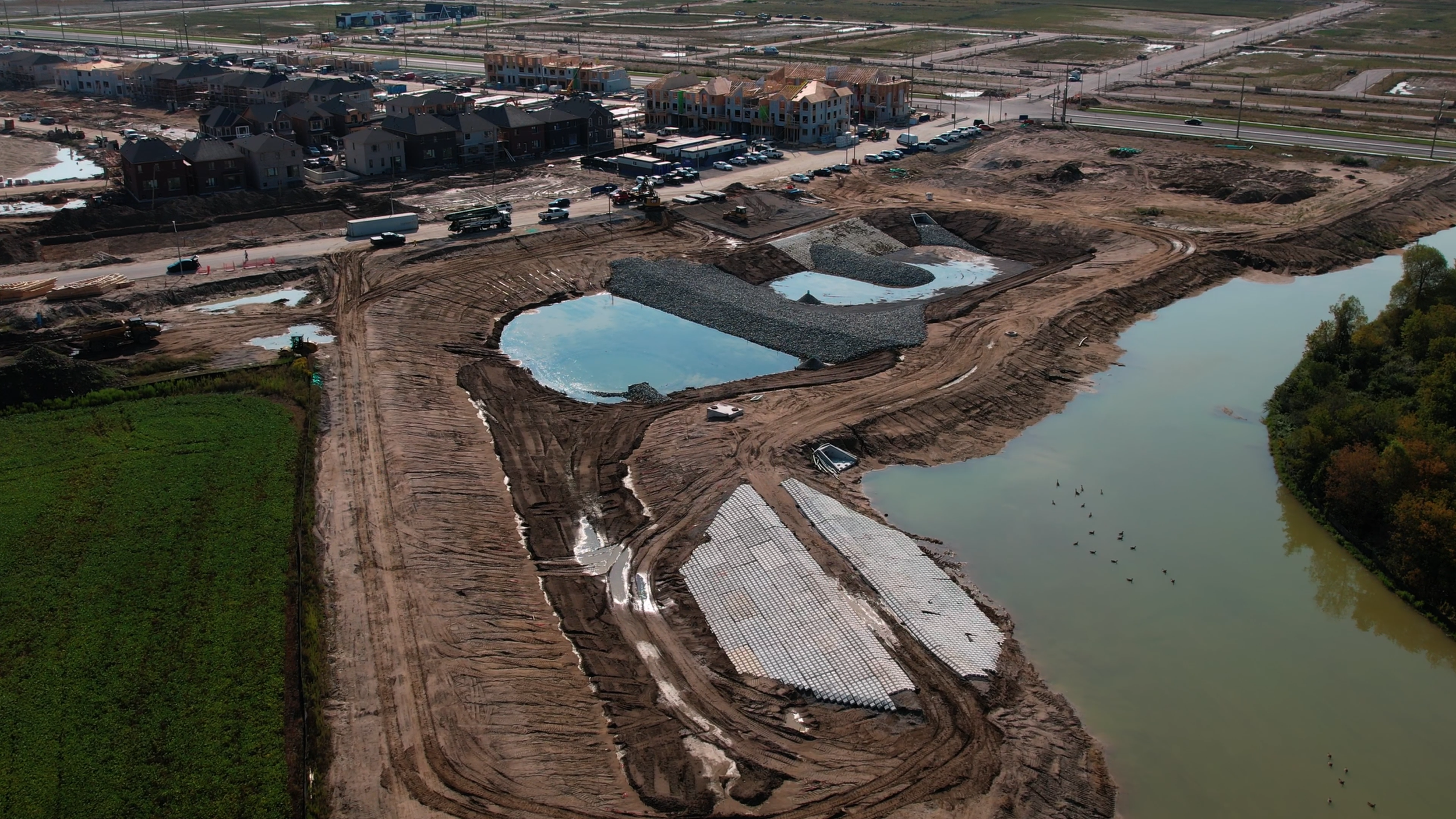 Aerial view of a construction site near a body of water, with piles of gravel, partially constructed buildings, and a green field.