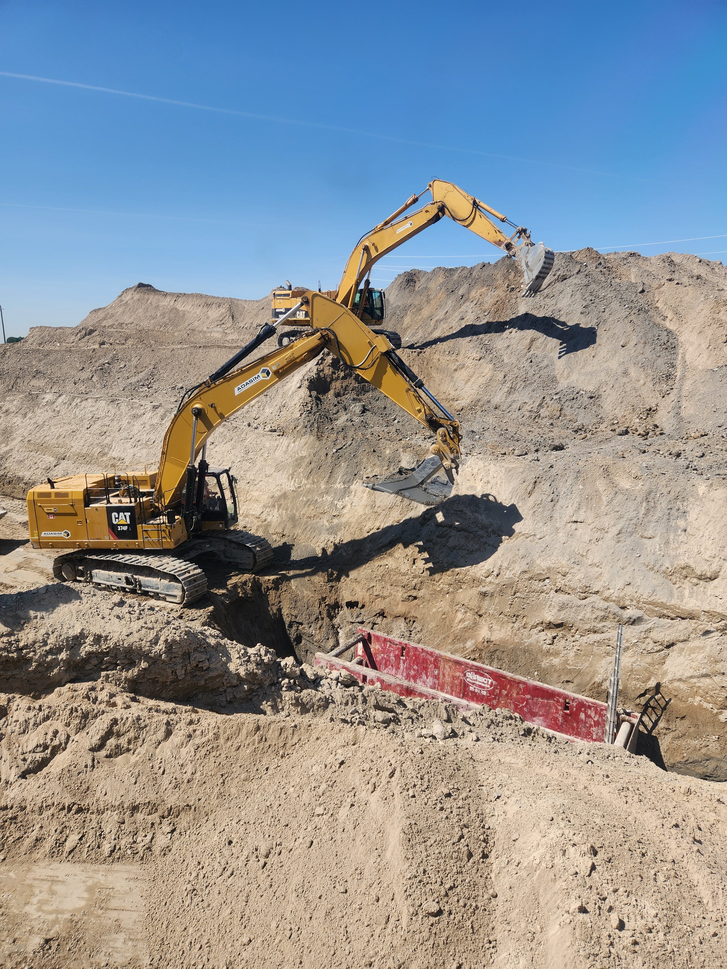 Two yellow excavators working on a construction site, moving dirt and earth, with clear blue sky in the background.