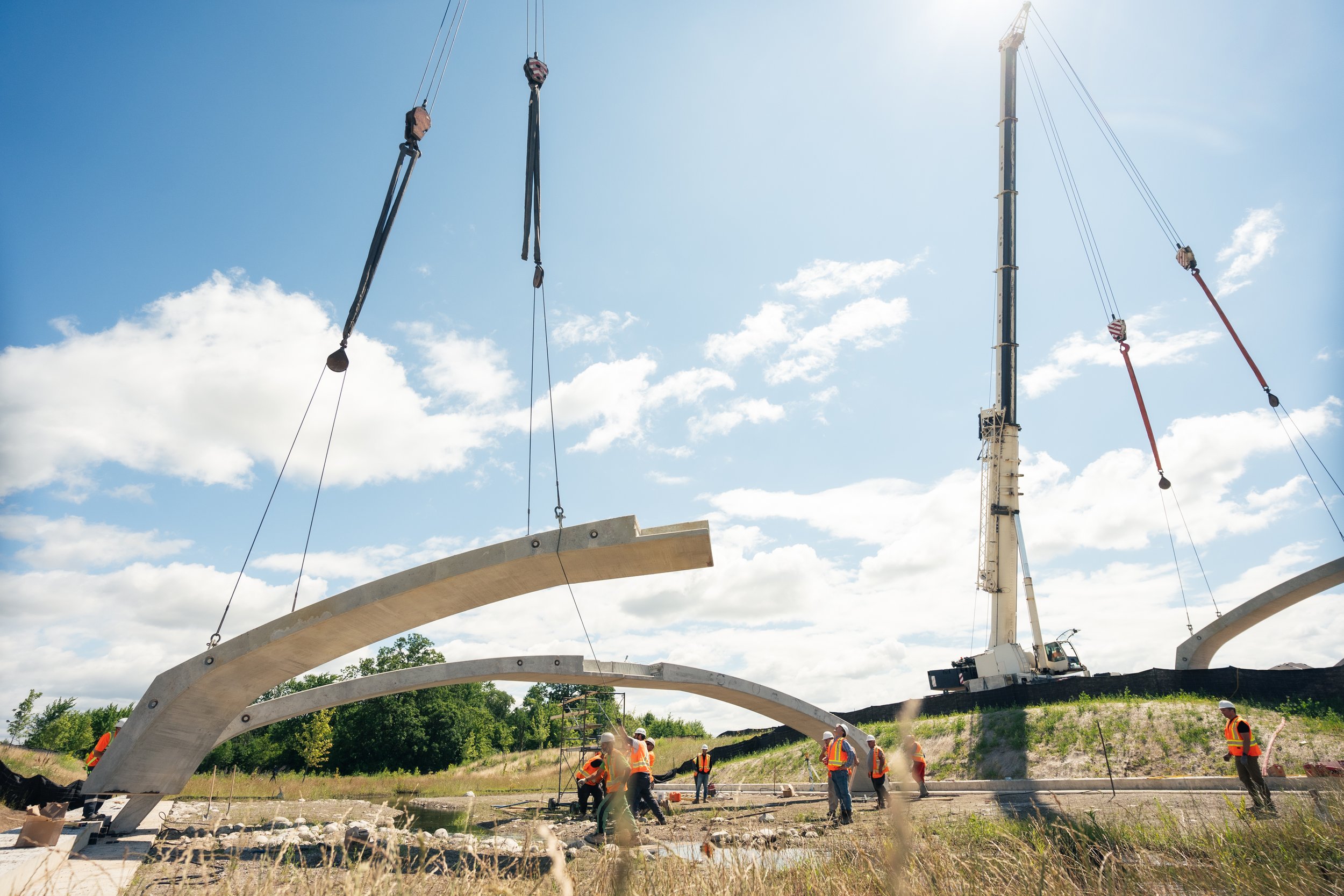 Construction workers in safety vests and helmets guiding large concrete arches into place with a crane on a construction site.