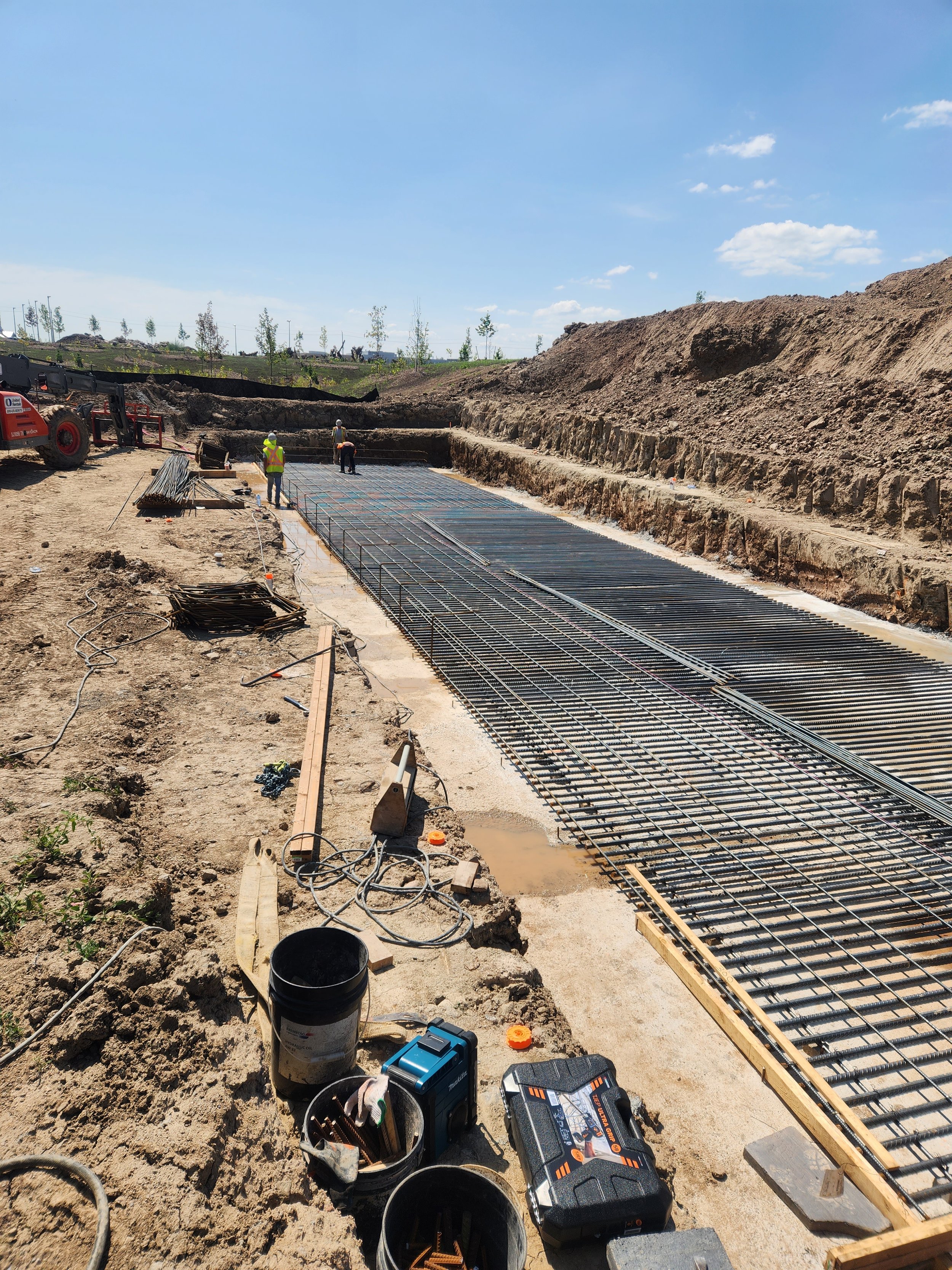 Construction workers working on a rebar foundation at a construction site with dirt and sky visible.