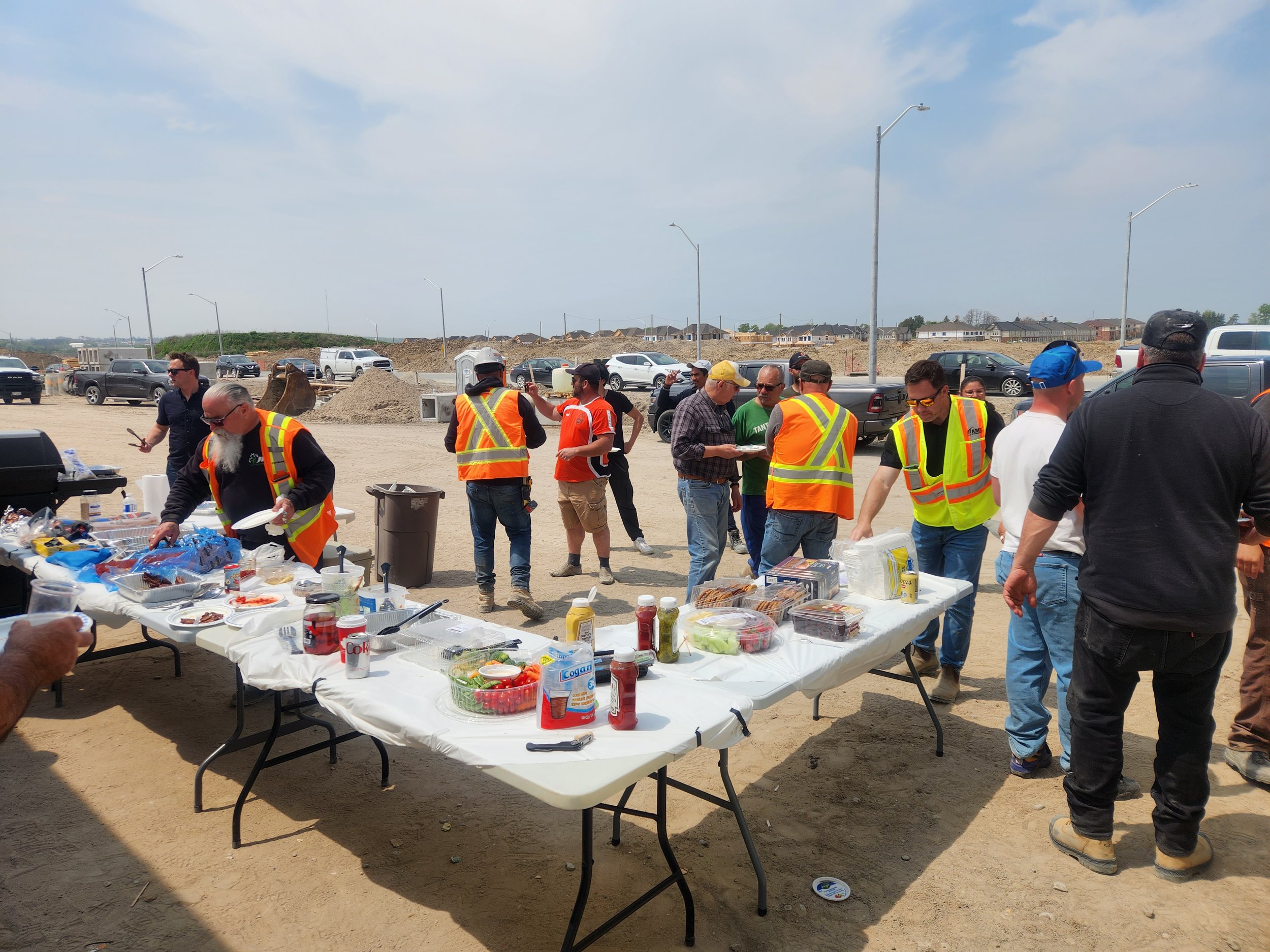 People gathered around tables with food during a construction site cookout, some wearing reflective safety vests, at a partly cloudy day.