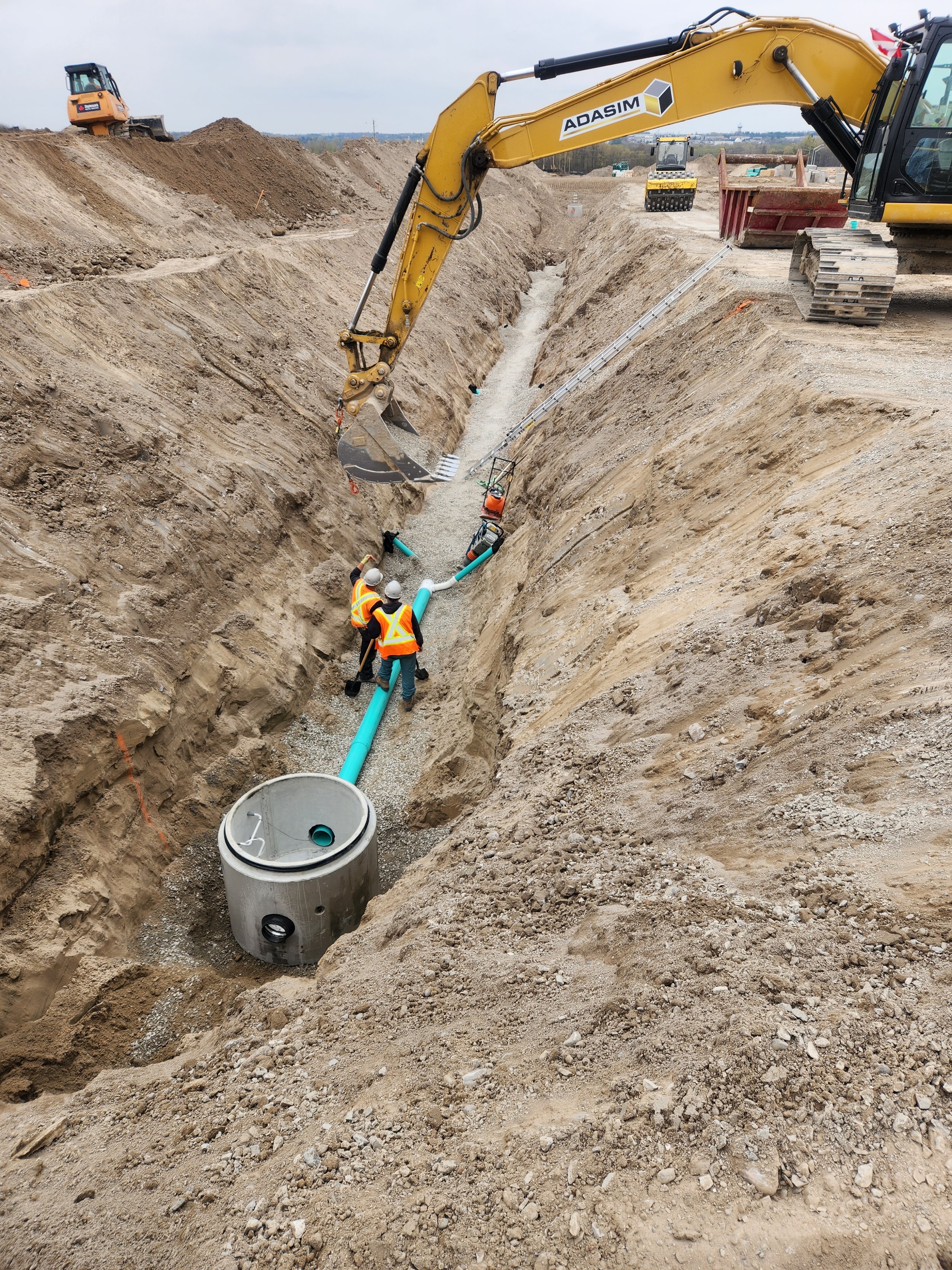 Construction workers installing large pipes in a deep trench with excavators on a dirt construction site.
