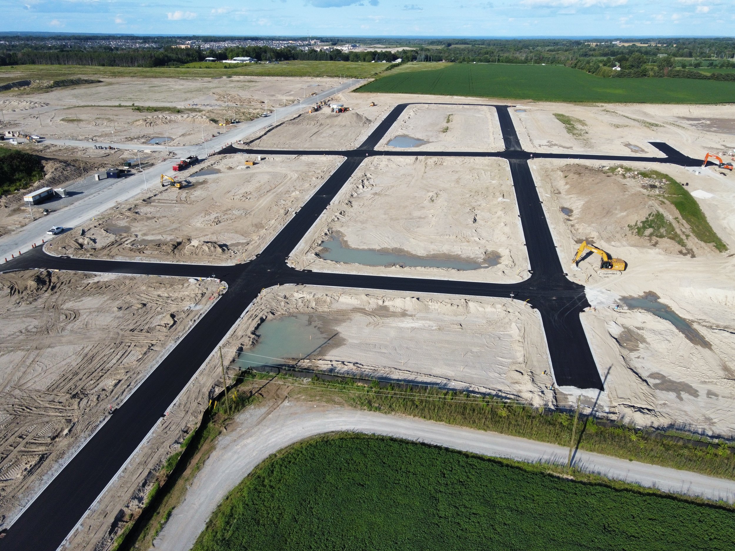 An aerial view of a construction site with freshly paved roads and several empty land plots, some with small water pools, and construction equipment.