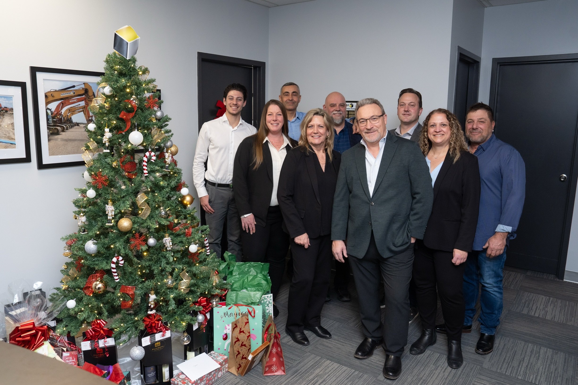 Group of nine people standing indoors near a decorated Christmas tree with presents underneath.