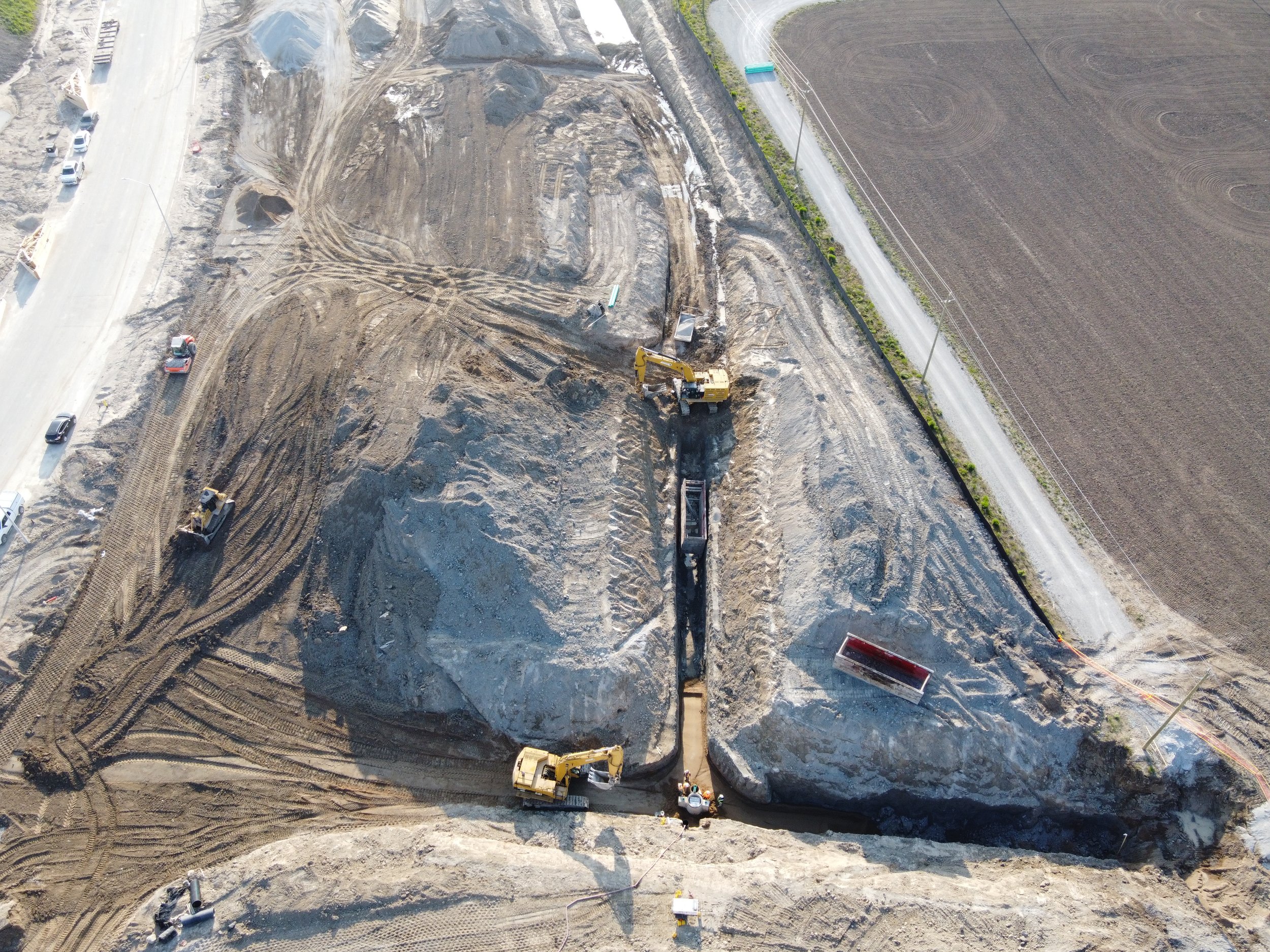 An aerial view of a construction site with heavy machinery digging into the ground, dirt roads, and surrounding farmland.