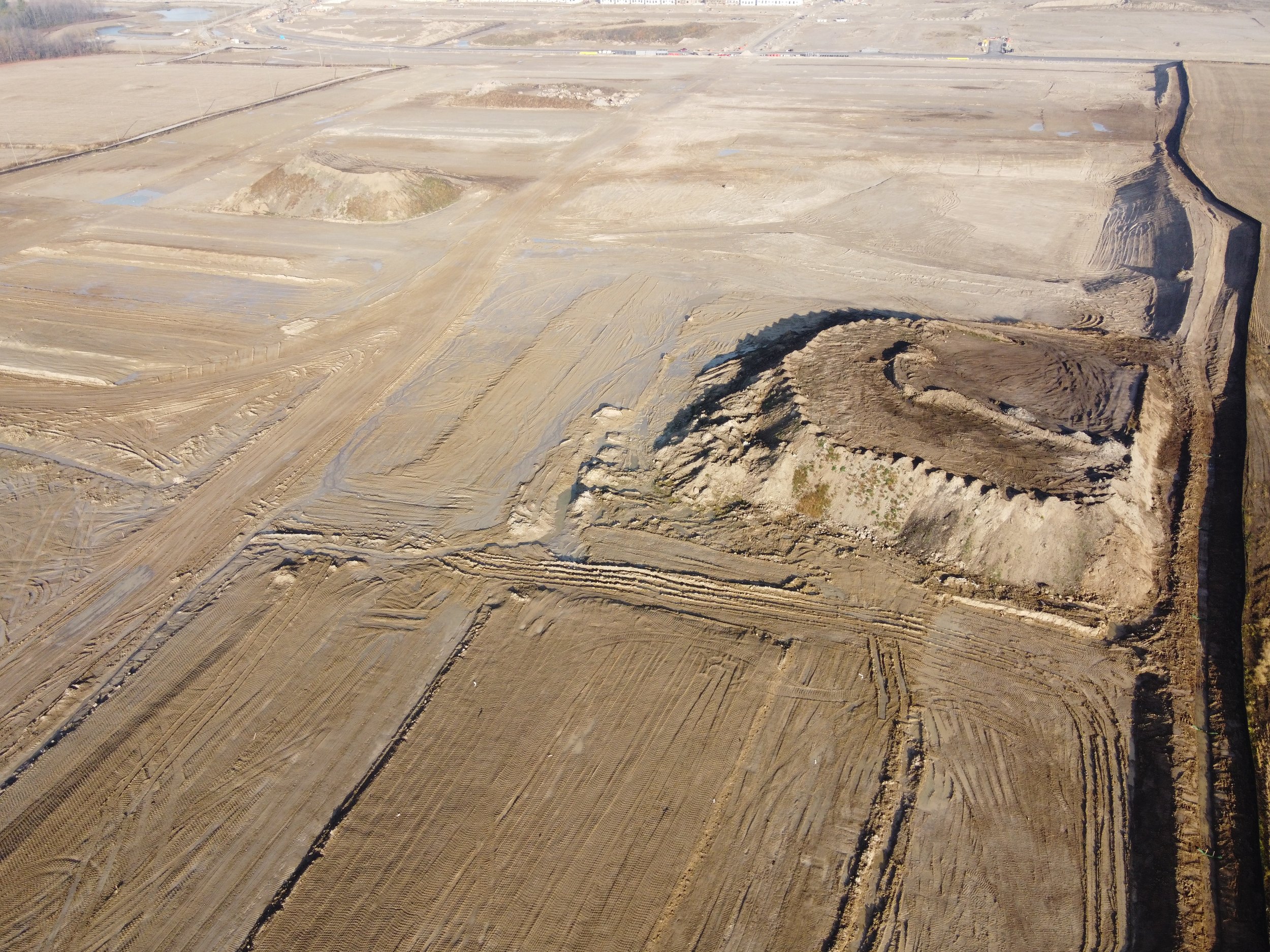 Aerial view of an expansive construction site with cleared land, dirt mounds, and areas prepared for development. The site is sectioned with visible dirt roads and barriers.
