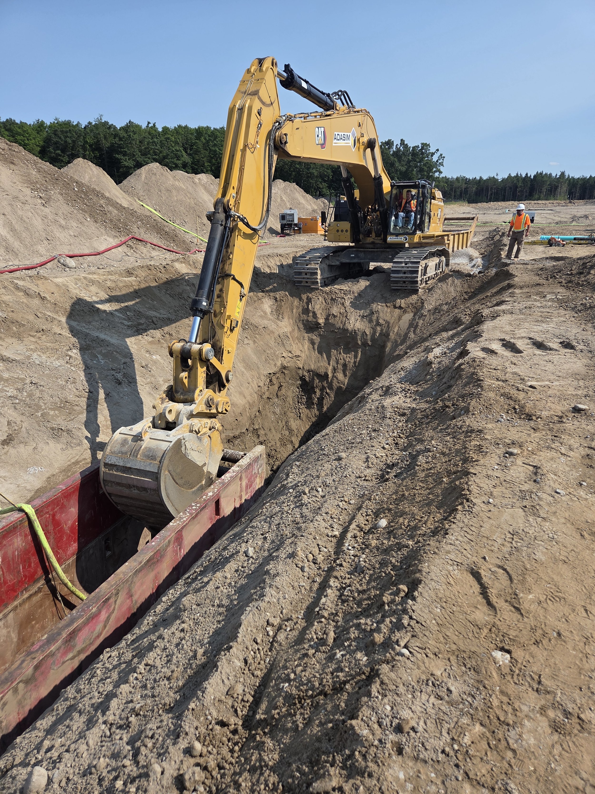 Construction site with a yellow excavator digging a trench, with workers and construction equipment in the background.