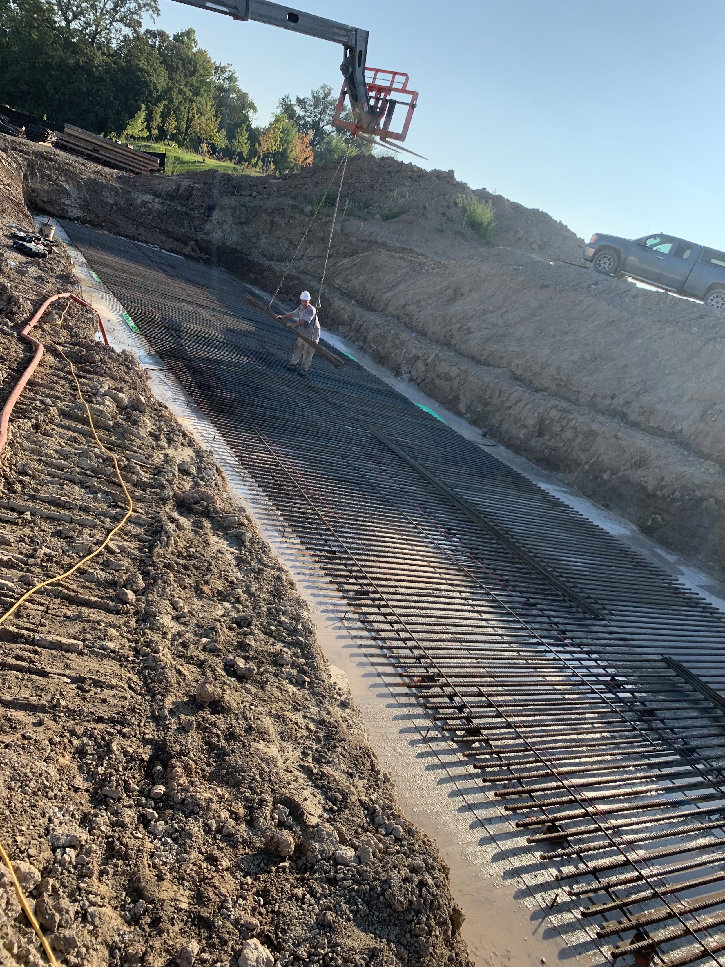 Construction workers installing reinforced steel bars in a highway or road under construction, with a crane overhead and a truck parked on the embankment.