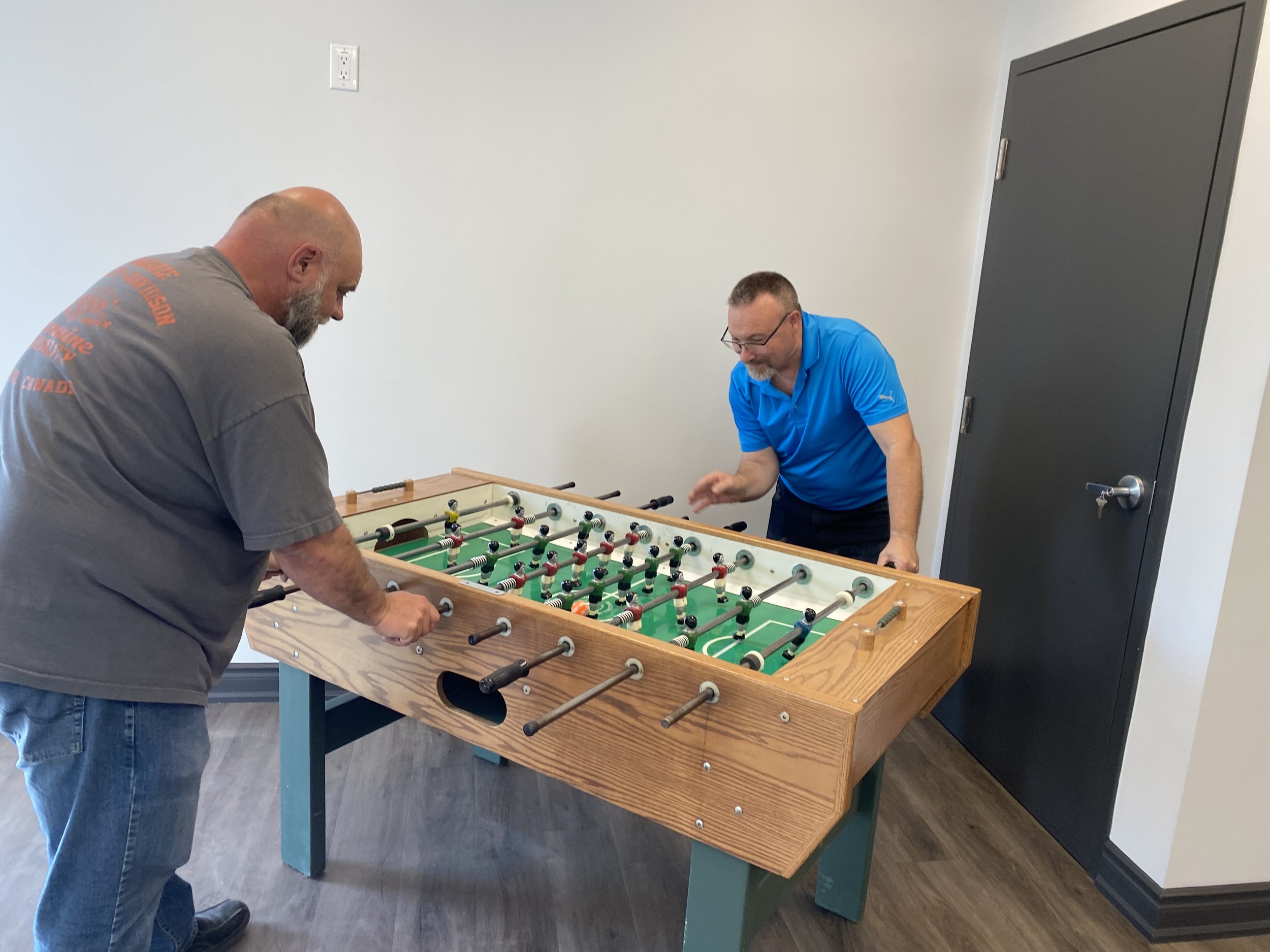 Two men playing foosball in a room with white walls and a gray door.