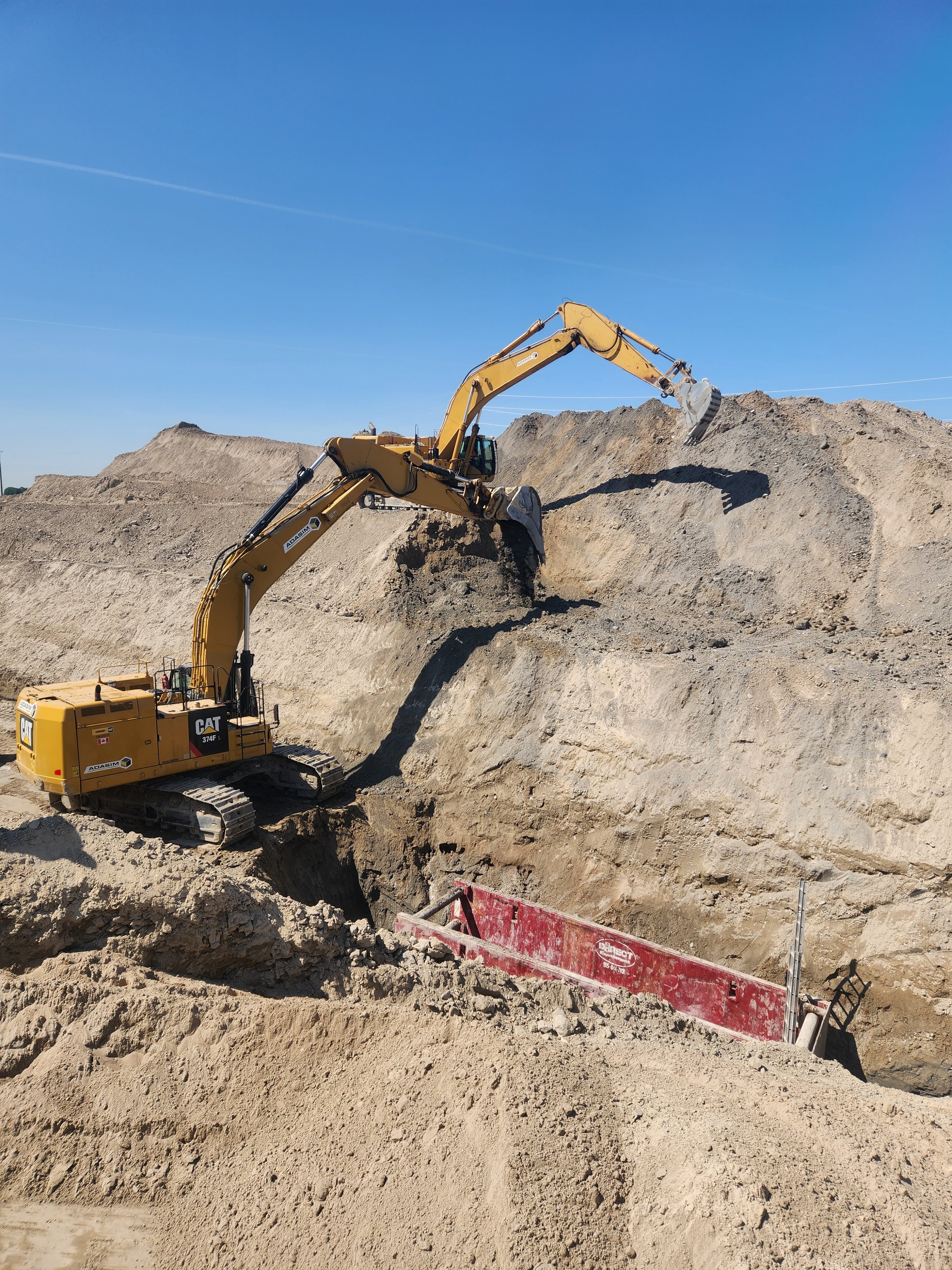 A construction site with two yellow excavators moving dirt and soil, with clear blue sky in the background.