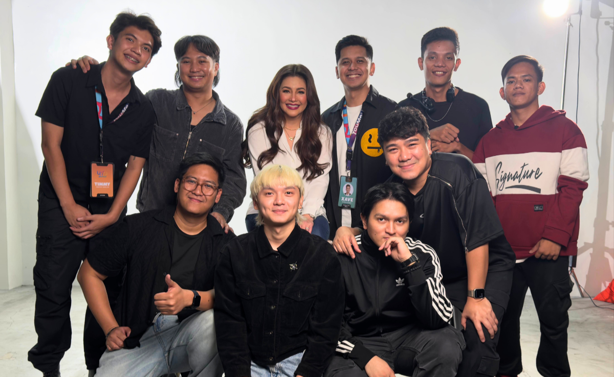 Group of ten young adults posing together for a photo in a studio setting, some wearing casual clothing and others with lanyards, smiling at the camera.