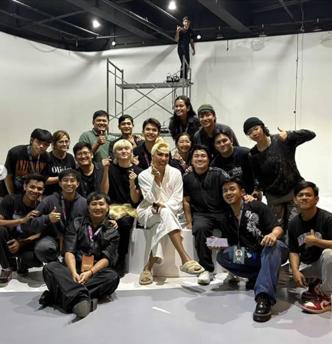 Group of diverse people gathered for a photo in a studio, with a woman in white sitting in the center, surrounded by smiling friends and a person standing on a scaffolding in the background.
