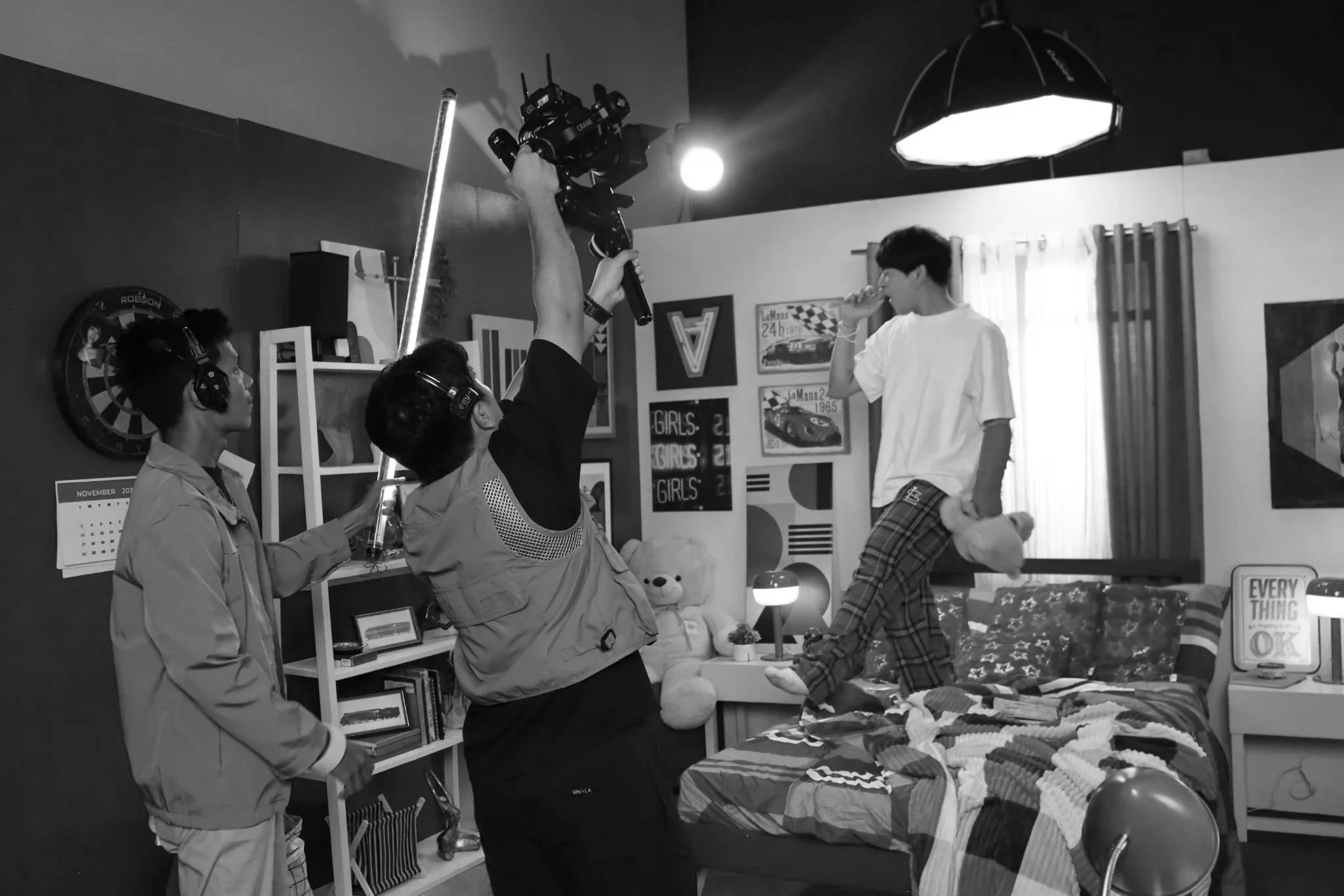 A film or television scene in a bedroom shows a young man on a bed holding a stuffed animal in one hand and a bong in the other, with crew members filming him with a professional camera. The room has posters, books, a teddy bear, and a lamp, and is decorated with curtains and artwork on the walls.