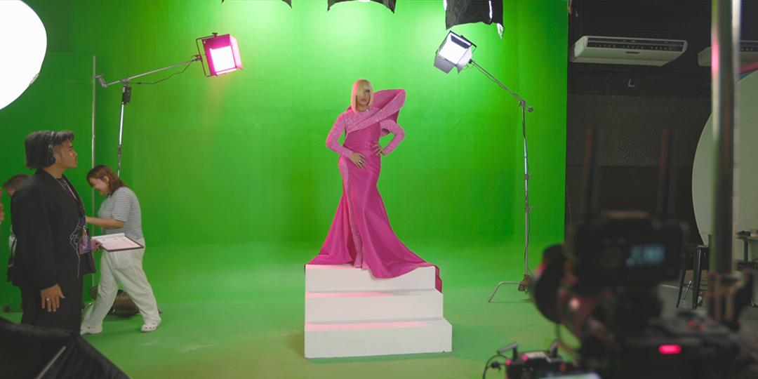 A woman in a bright pink gown with dramatic shoulder accents posing on a platform in front of a green screen studio, with crew members and lighting equipment visible.