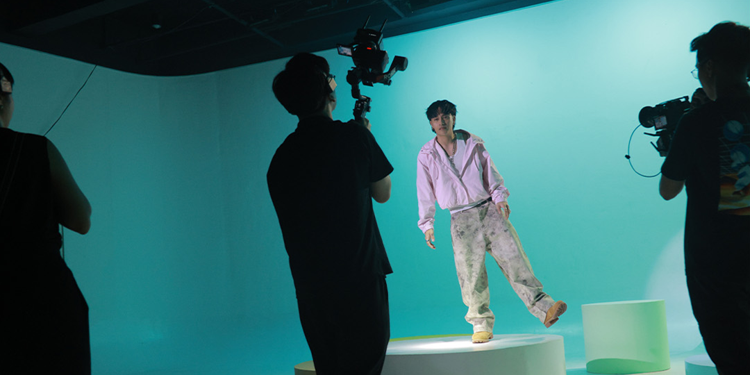 A person in casual clothing standing on a raised platform during a photoshoot or video shoot, with a teal backdrop and crew members filming.