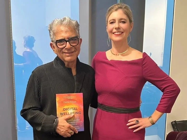 An older man (Deepak Chopra) with gray hair, glasses, and a black striped shirt holding a book titled 'Digital Wellbeing,' standing next to Caitlin Krause in a magenta dress, both smiling indoors with glass windows in the background.
