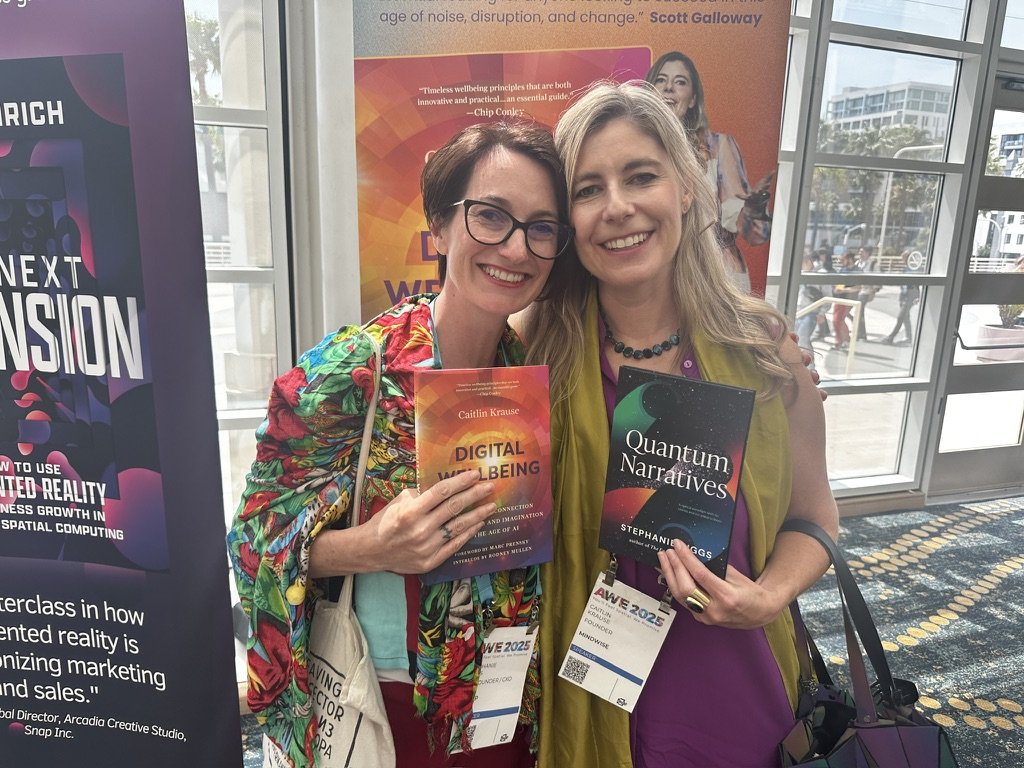 Two women (Caitlin Krause, author, keynote speaker)  smiling and holding books at a conference, standing in front of a promotional poster and glass windows.