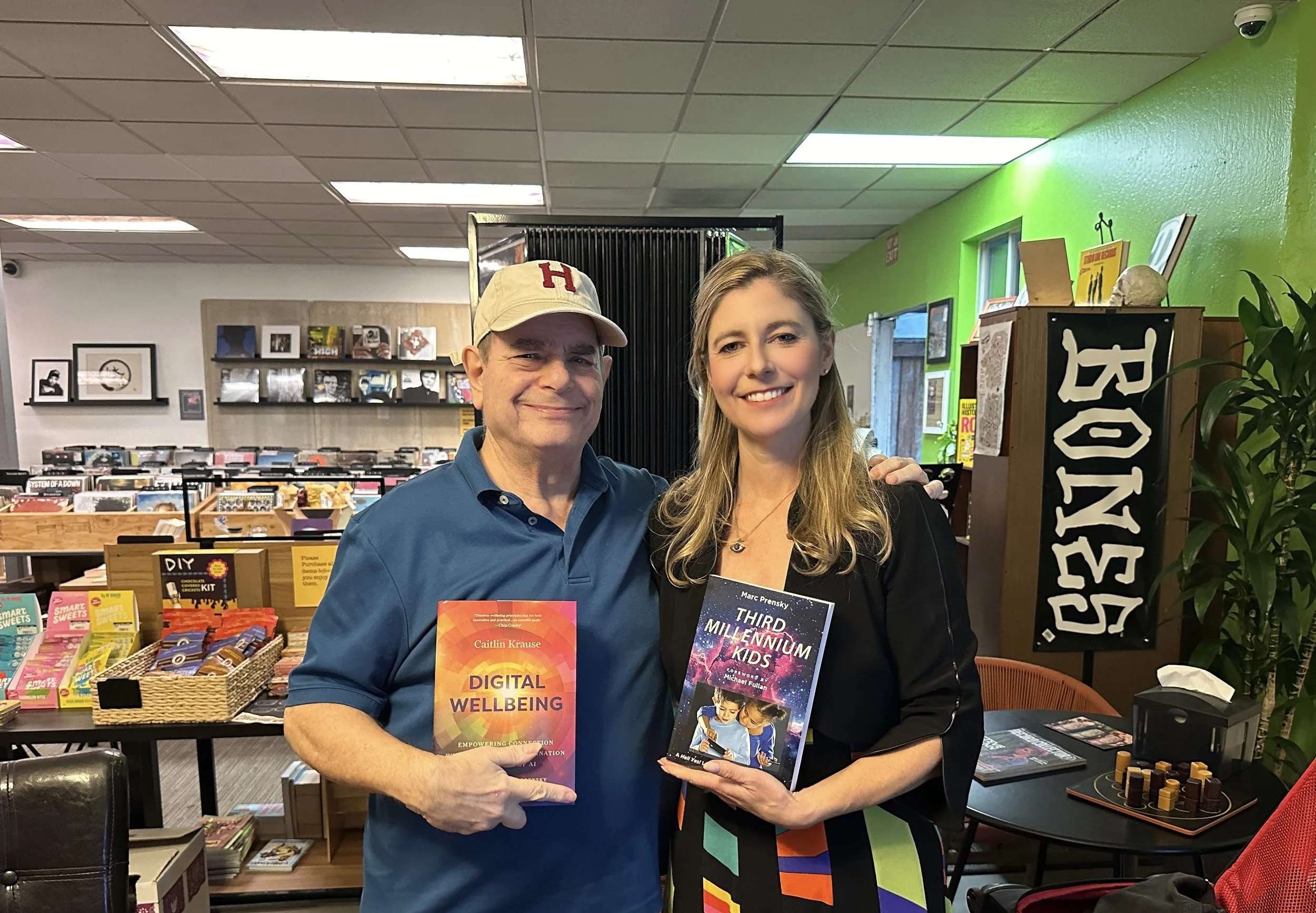 A man and woman (Caitlin Krause, author, keynote speaker) holding books inside a bookstore, standing in front of bookshelves, smiling at the camera.