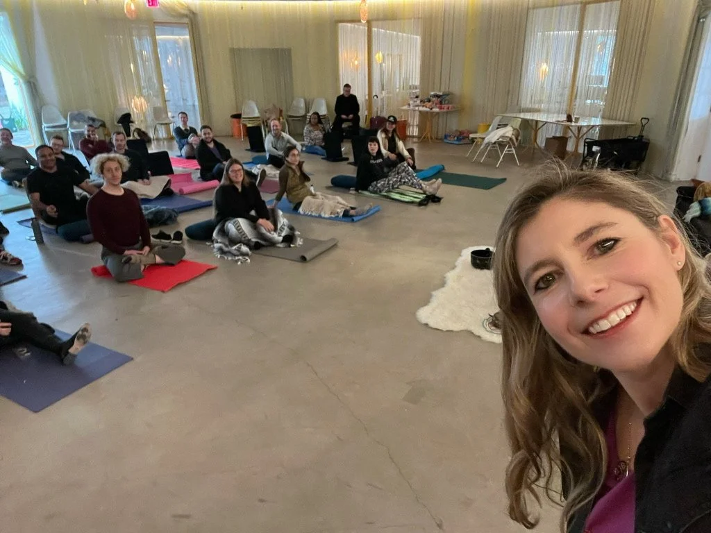 A group of people seated on yoga mats in a spacious, well-lit room, participating in a yoga or wellness class, with a woman taking a selfie at the front.