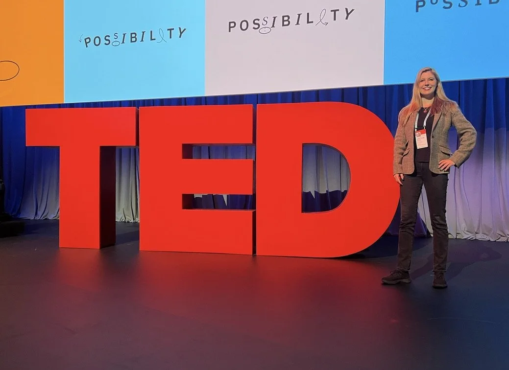 (Caitlin Krause, author, keynote speaker)  standing next to large red letters spelling 'TED' on a stage at a conference, with a screen in the background displaying the word 'POSSIBILITY' in different styles.