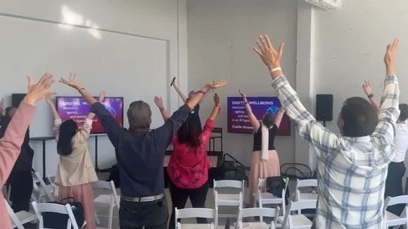 People standing with arms raised in a classroom or conference room during a workshop or presentation.
