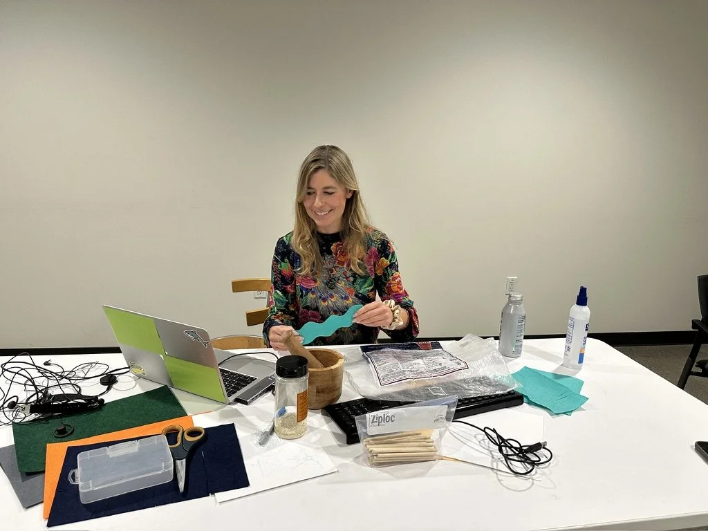 A woman (Caitlin Krause) keynote speaker, with long blonde hair sitting at a white table outside, smiling as she works on a turquoise-colored craft project with various crafting supplies around her, including scissors, a laptop, and glue.