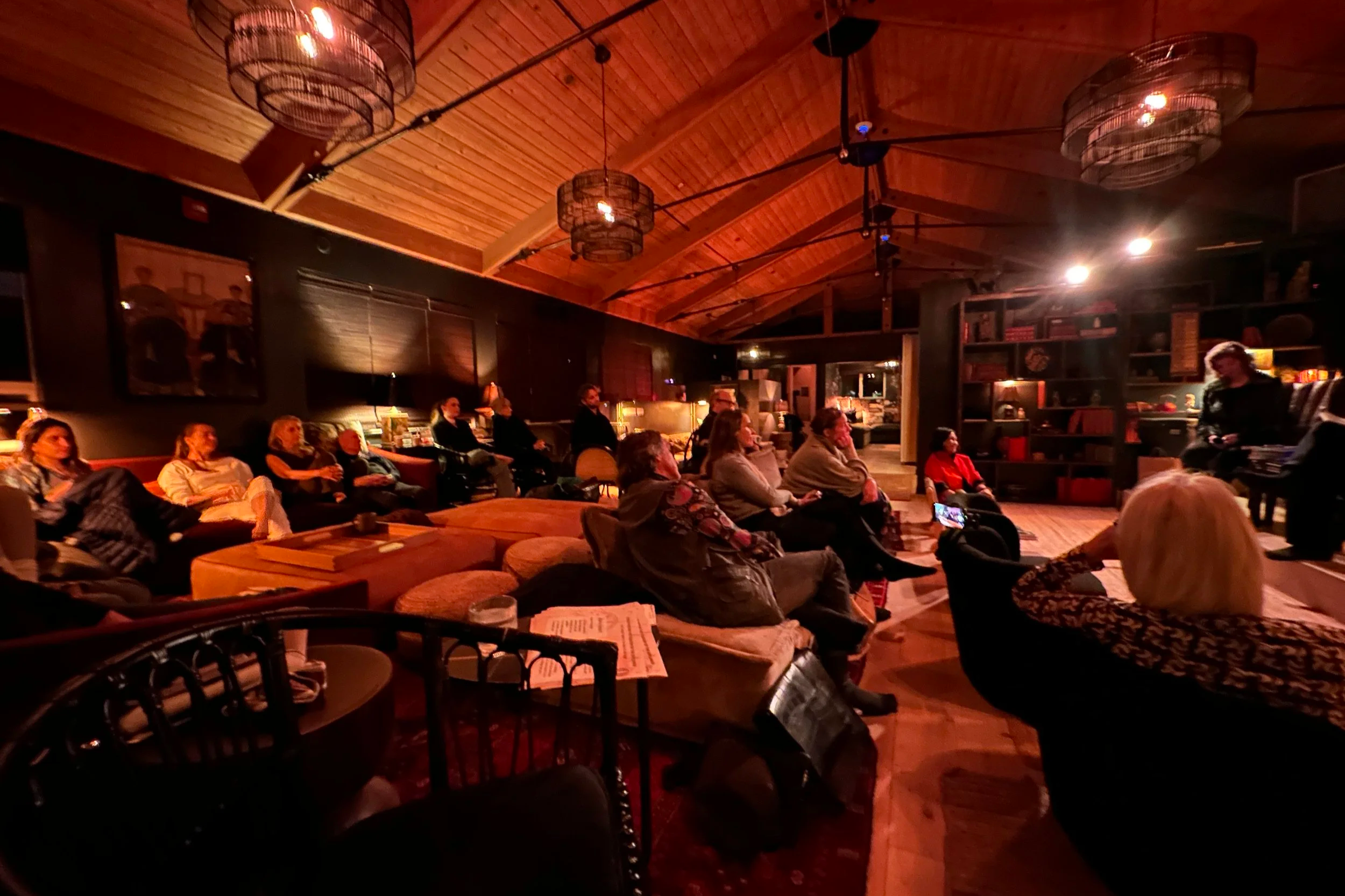 People seated and watching a live performance or presentation in a cozy, dimly lit room with wooden walls and ceiling, decorated with hanging light fixtures.