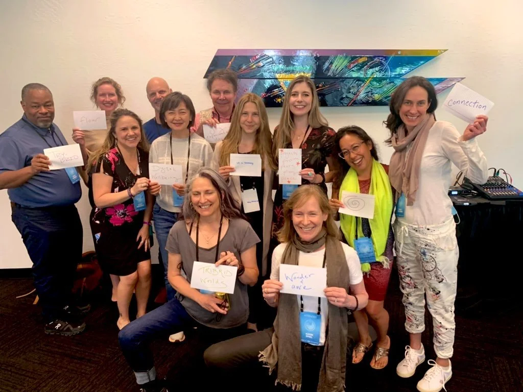 Group of people smiling at a conference or workshop, holding up signs with words like 'flow,' 'connection,' and 'Tribu,' with a colorful artwork on the wall behind them.